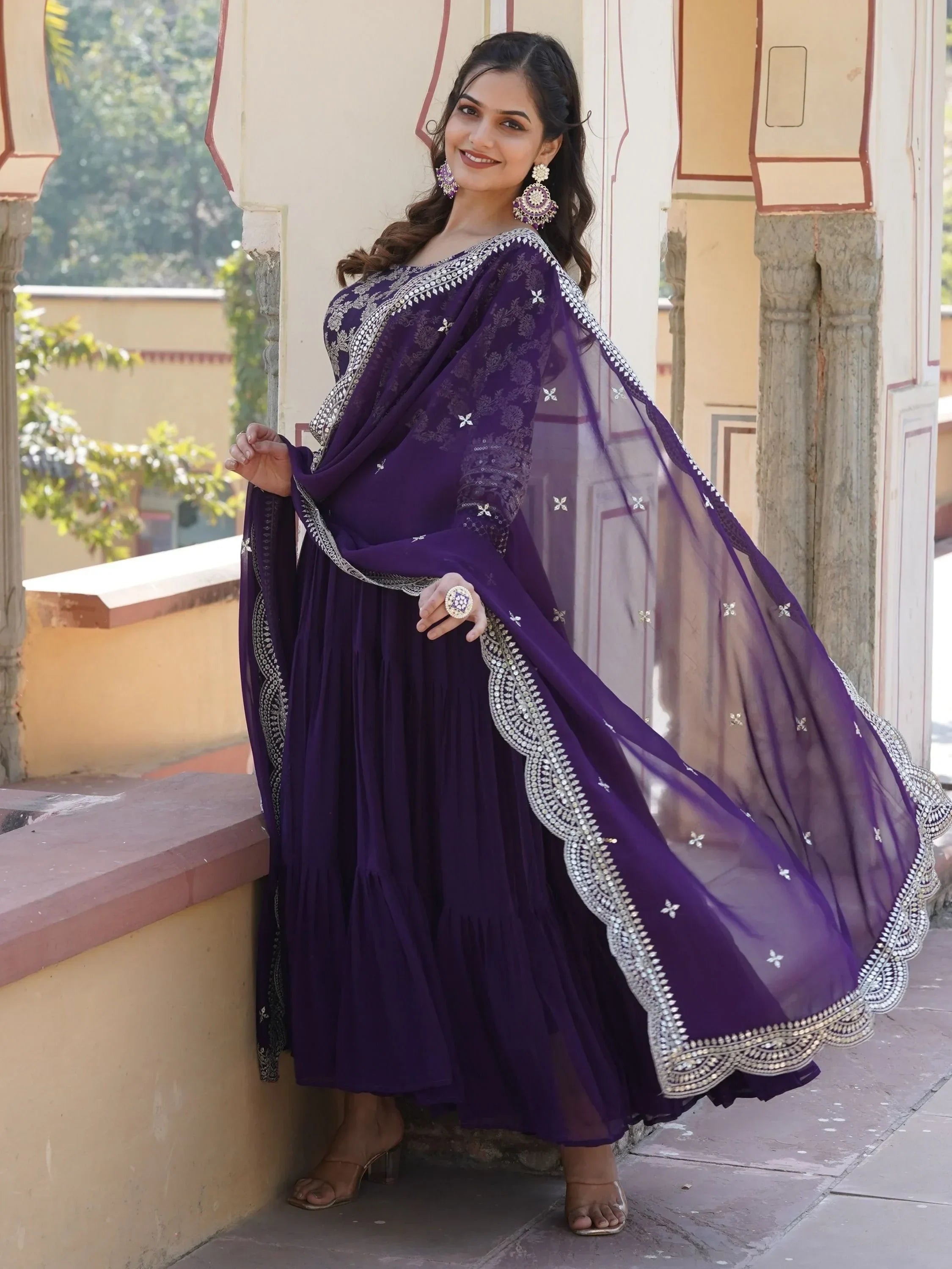 Woman wearing a purple embroidered Indian gown with dupatta, ethnic jewelry, standing outdoors