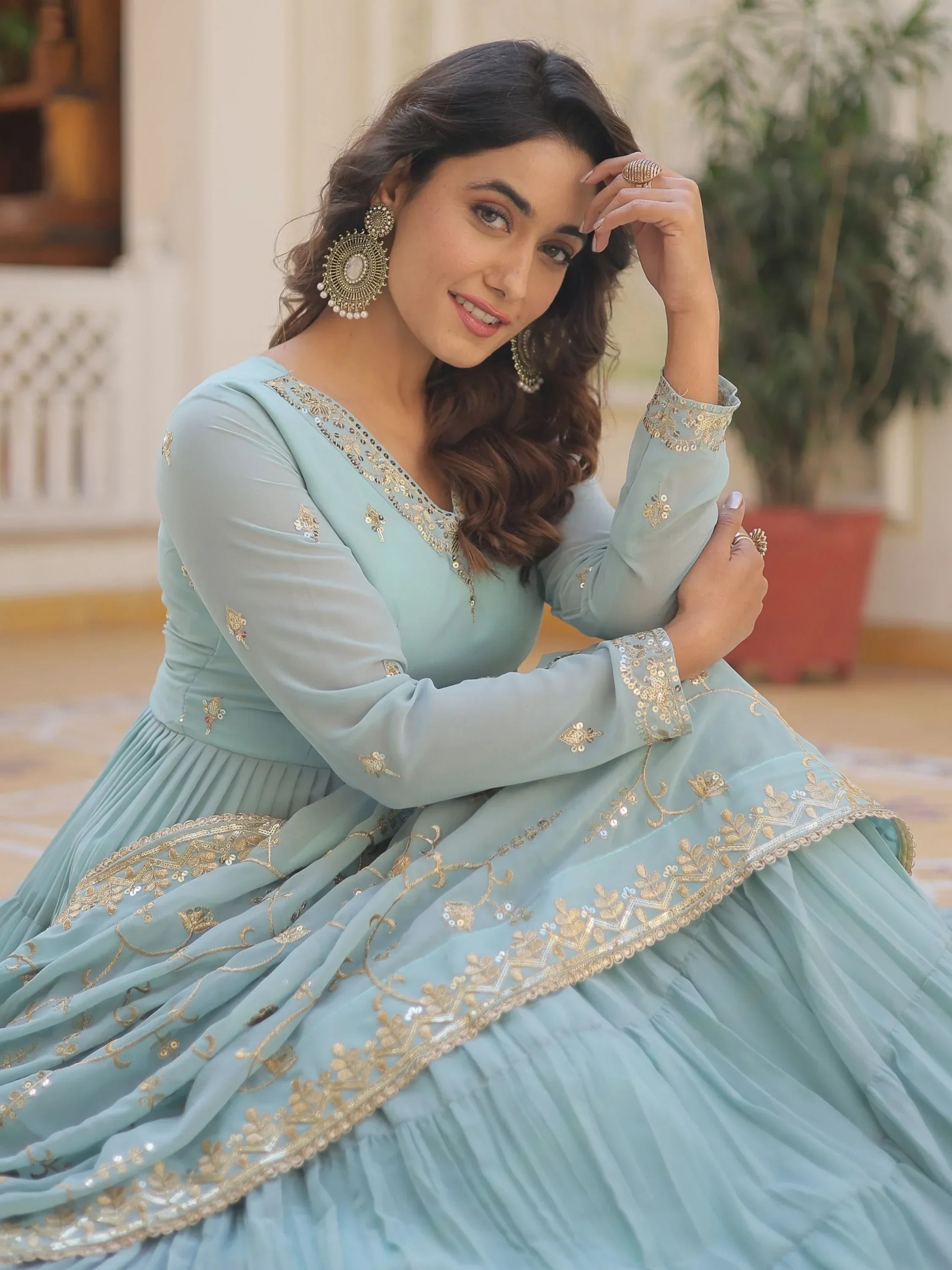 Woman in pastel blue Indian ethnic gown with gold embroidery, sitting indoors, smiling