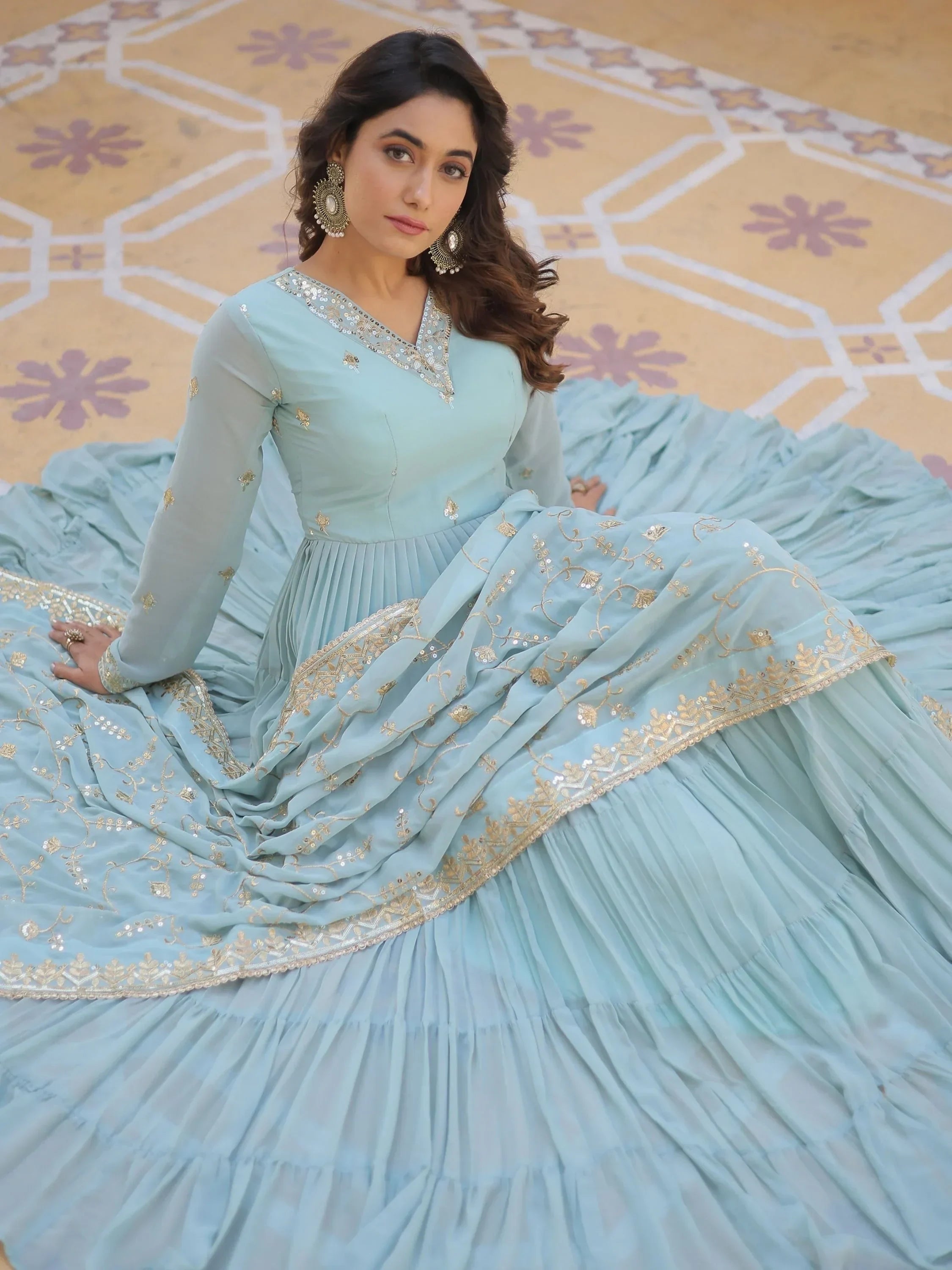 Woman in light blue embroidered Anarkali gown with gold details, seated on patterned floor