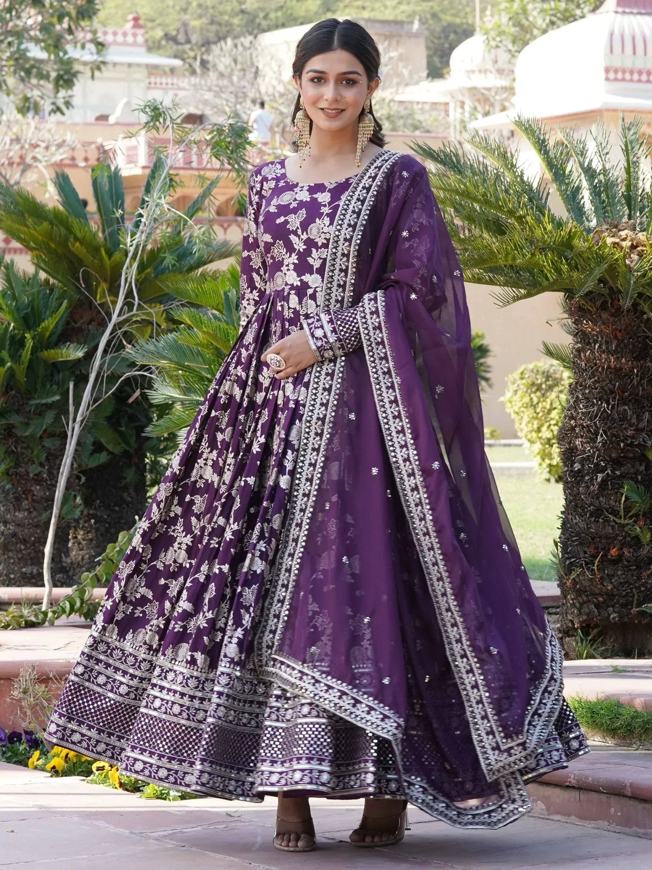 Woman in a purple embroidered ethnic gown with dupatta, standing outdoors by greenery