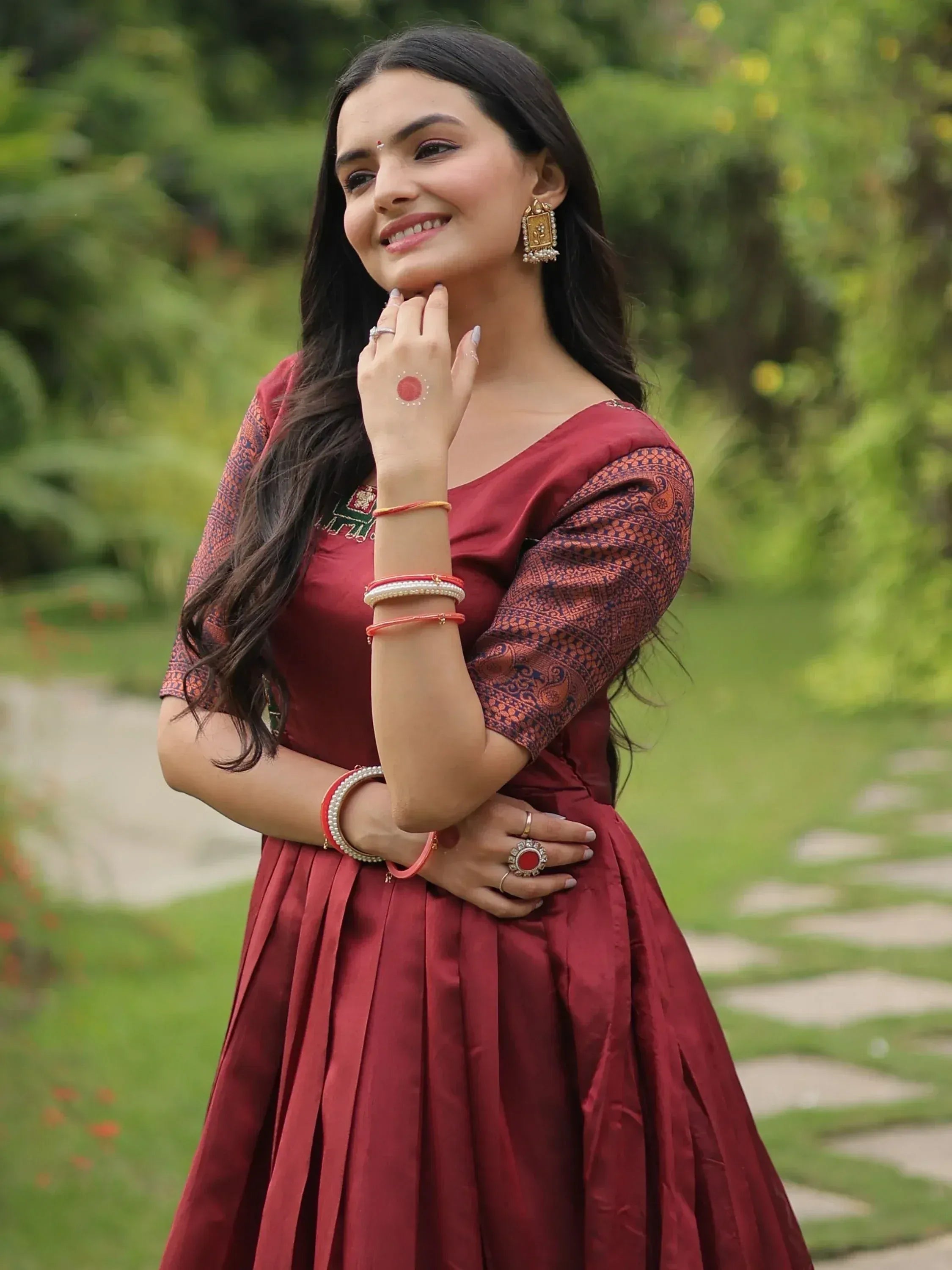 Woman in maroon ethnic gown with printed sleeves, traditional jewelry, smiling outdoors