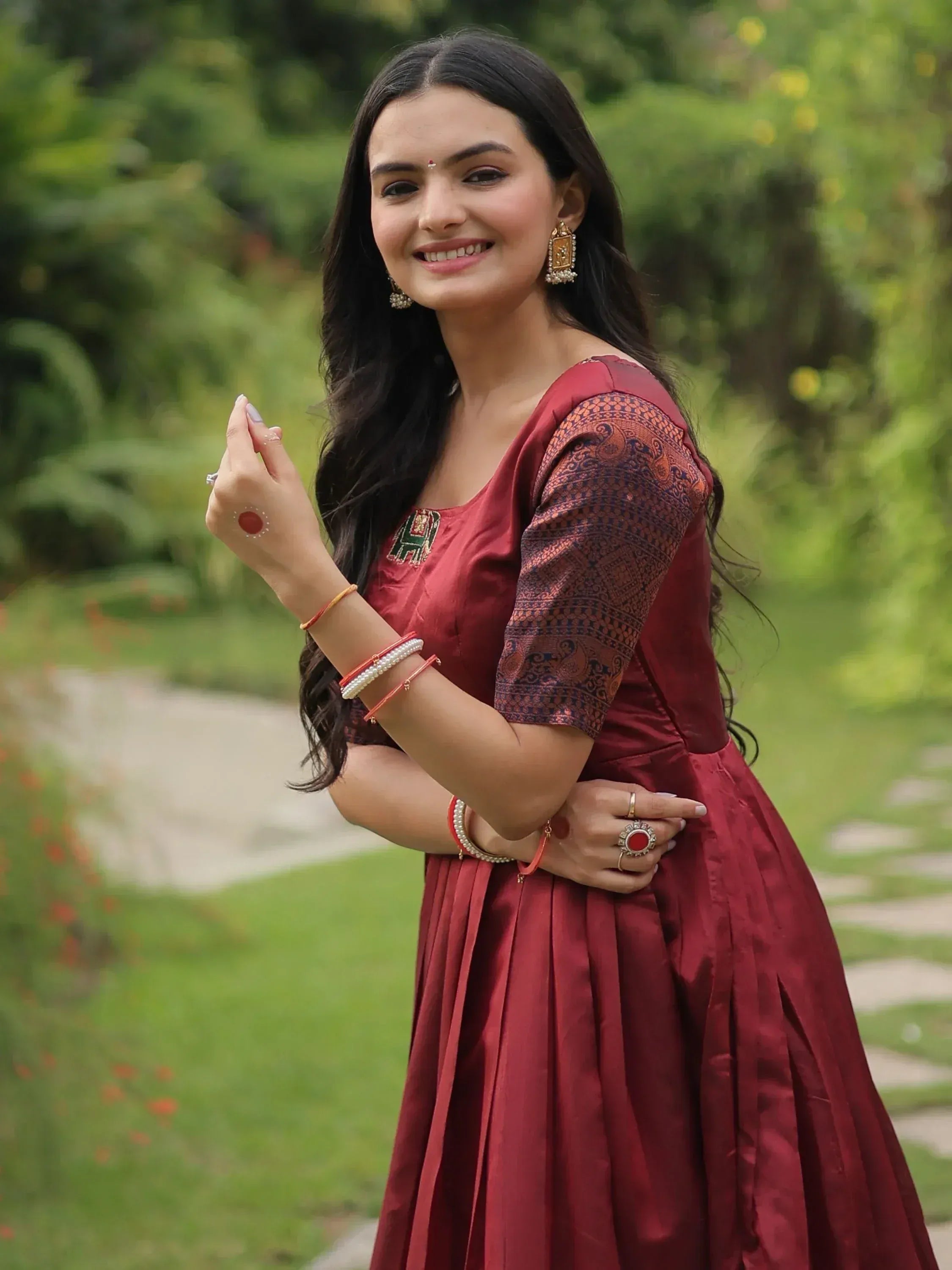 Woman in maroon ethnic gown with printed sleeves, traditional jewelry, smiling outdoors