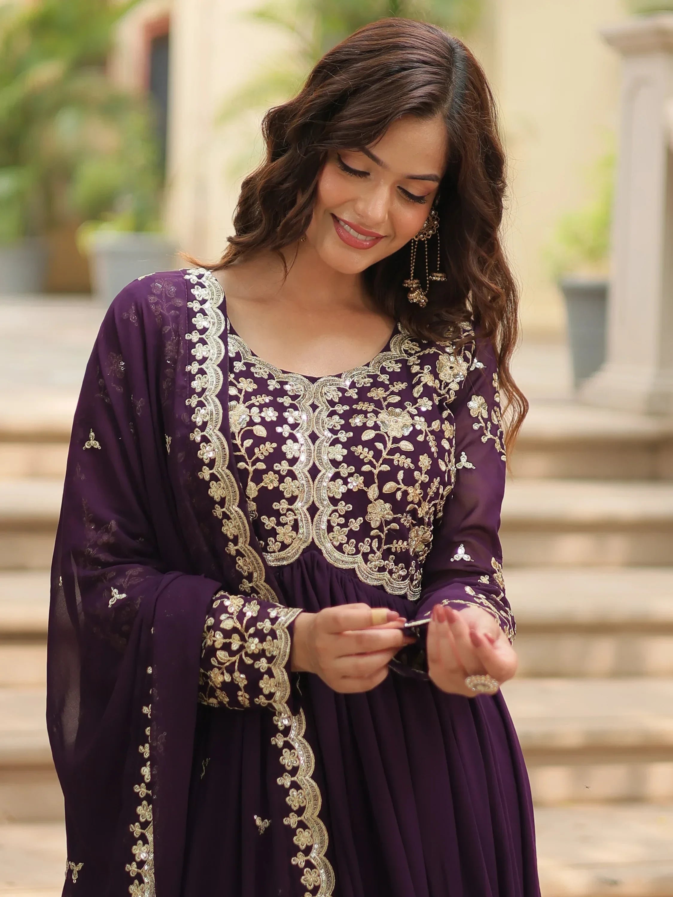 Woman in purple and gold embroidered ethnic gown, standing outdoors, smiling