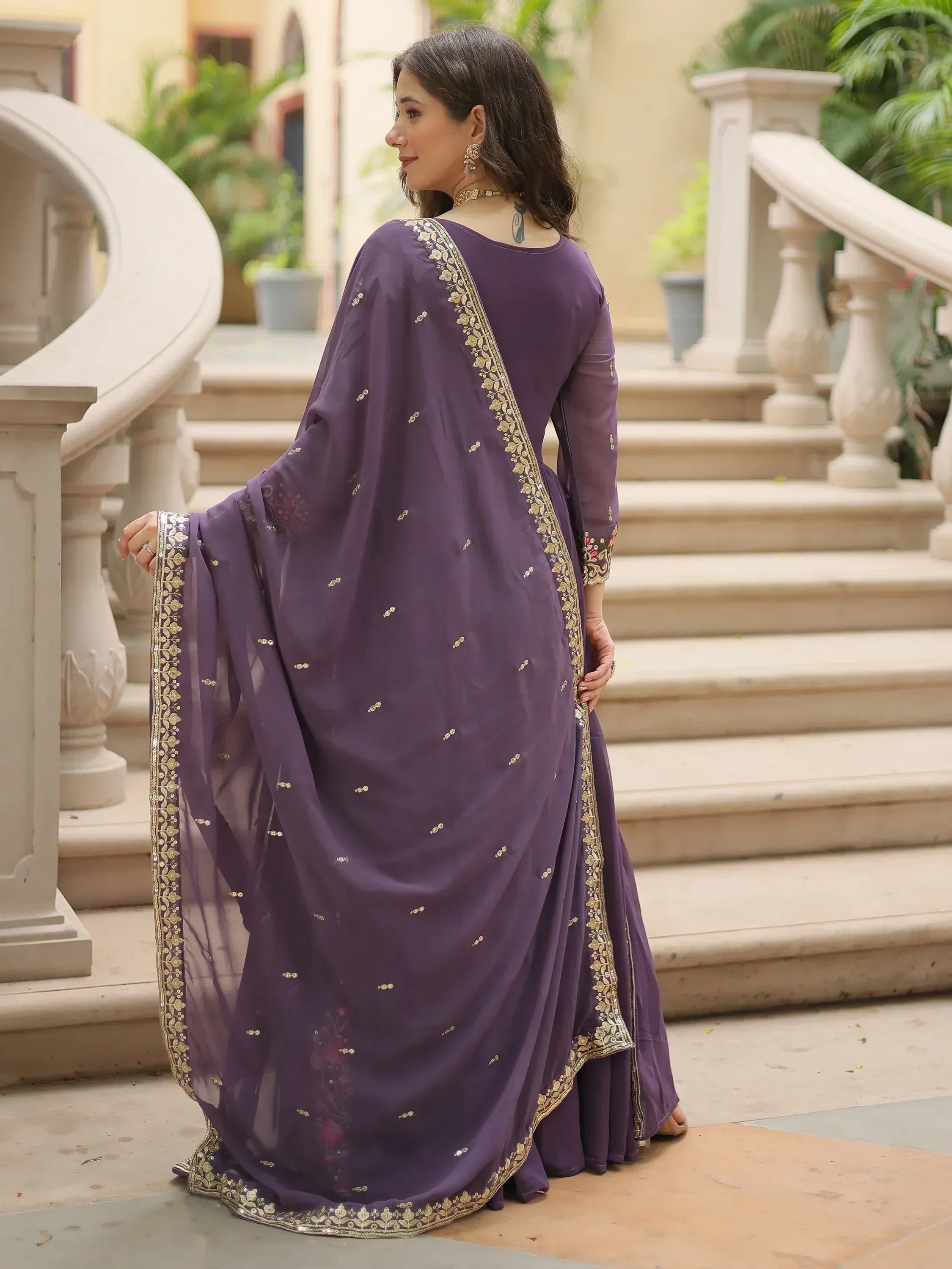 Woman in a purple ethnic saree with gold border, standing by a staircase outdoors