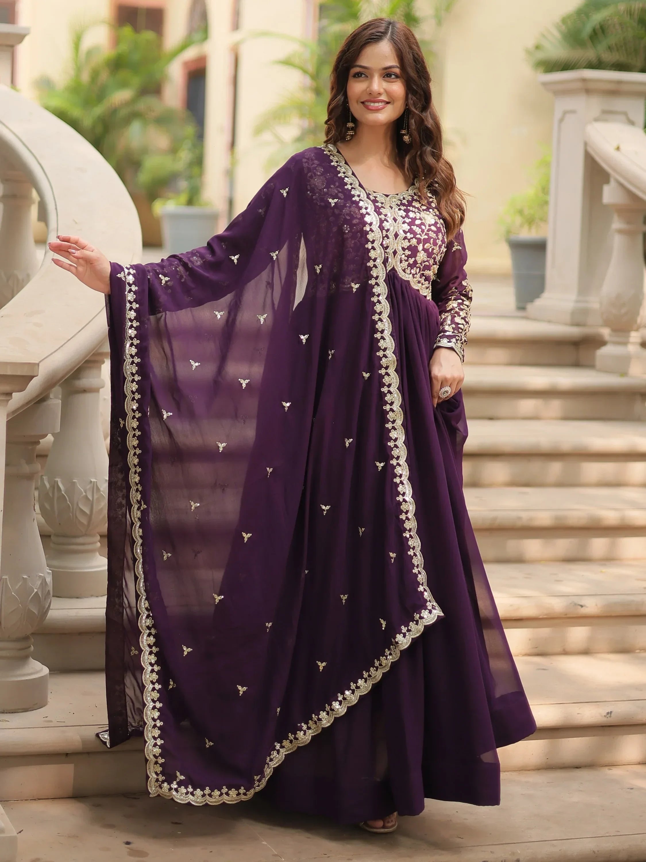 Woman wearing a purple embroidered Anarkali gown with dupatta, posing outdoors on steps