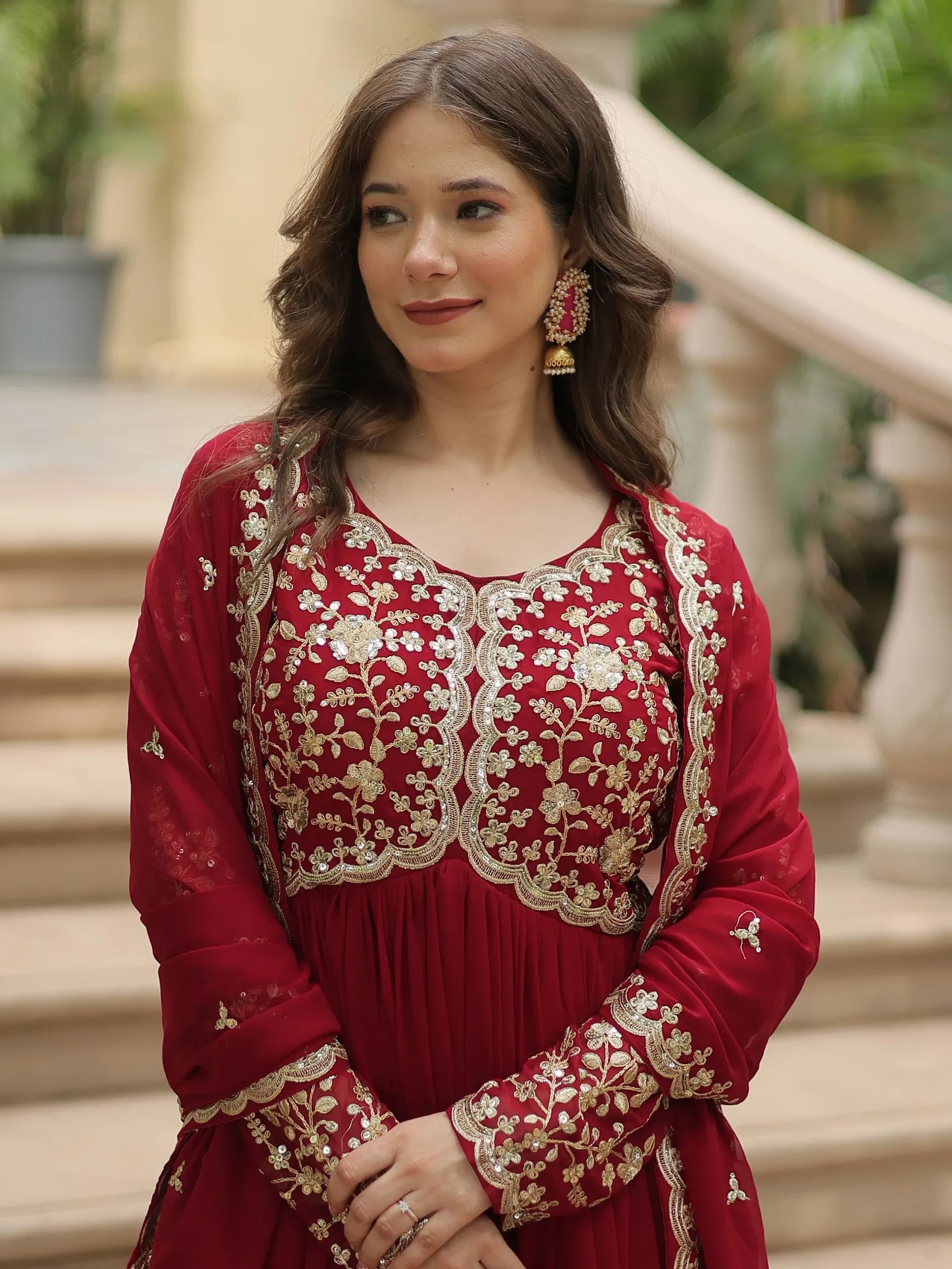 Woman wearing a red ethnic gown with gold embroidery, traditional earrings, outdoors