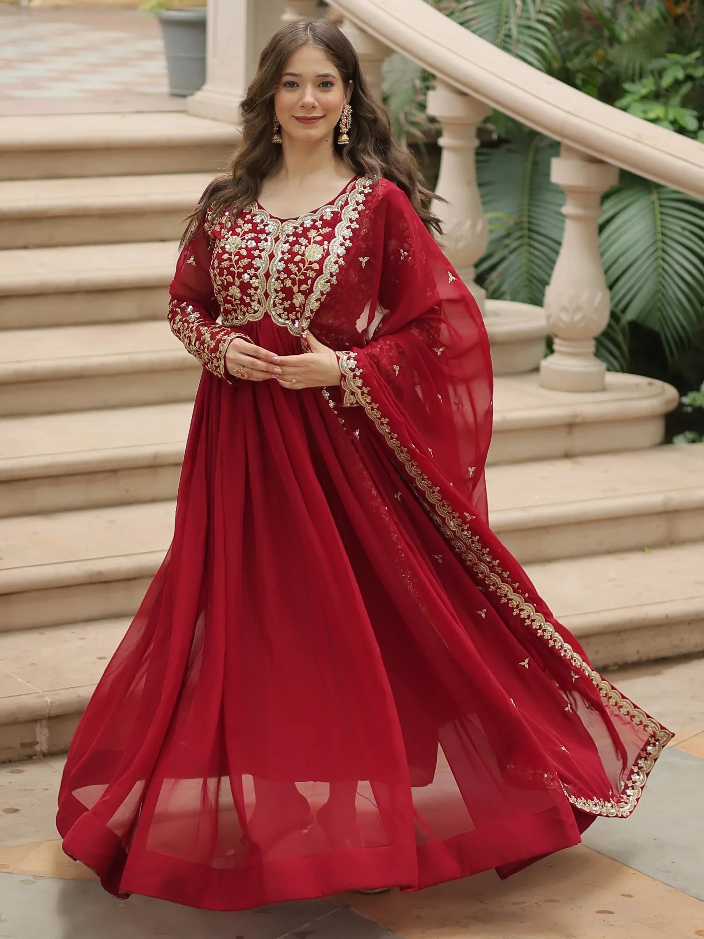 Woman in a red embroidered Anarkali suit with gold details, posing outdoors on stairs