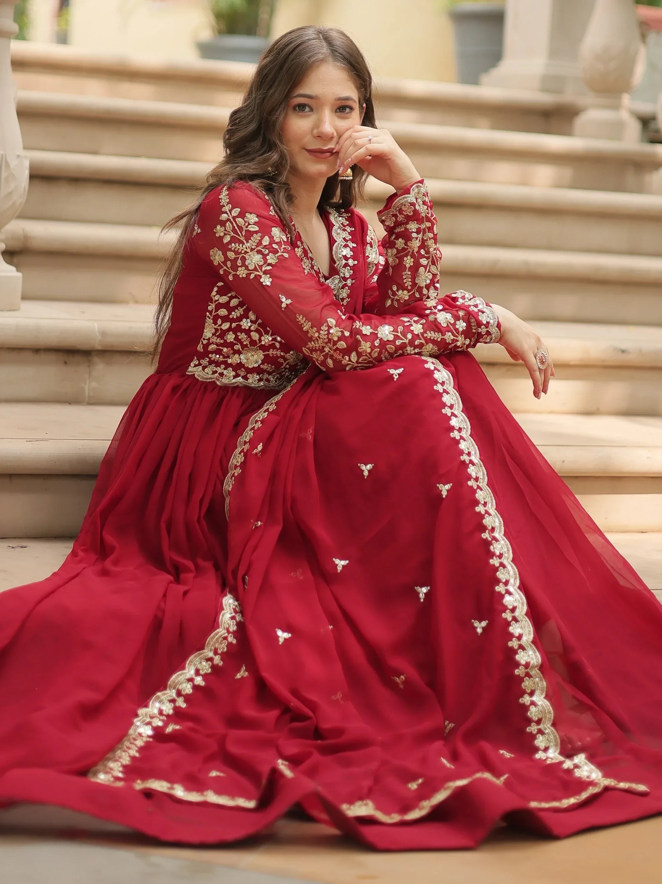 Woman in red embroidered lehenga gown with gold floral details, sitting on steps outdoors
