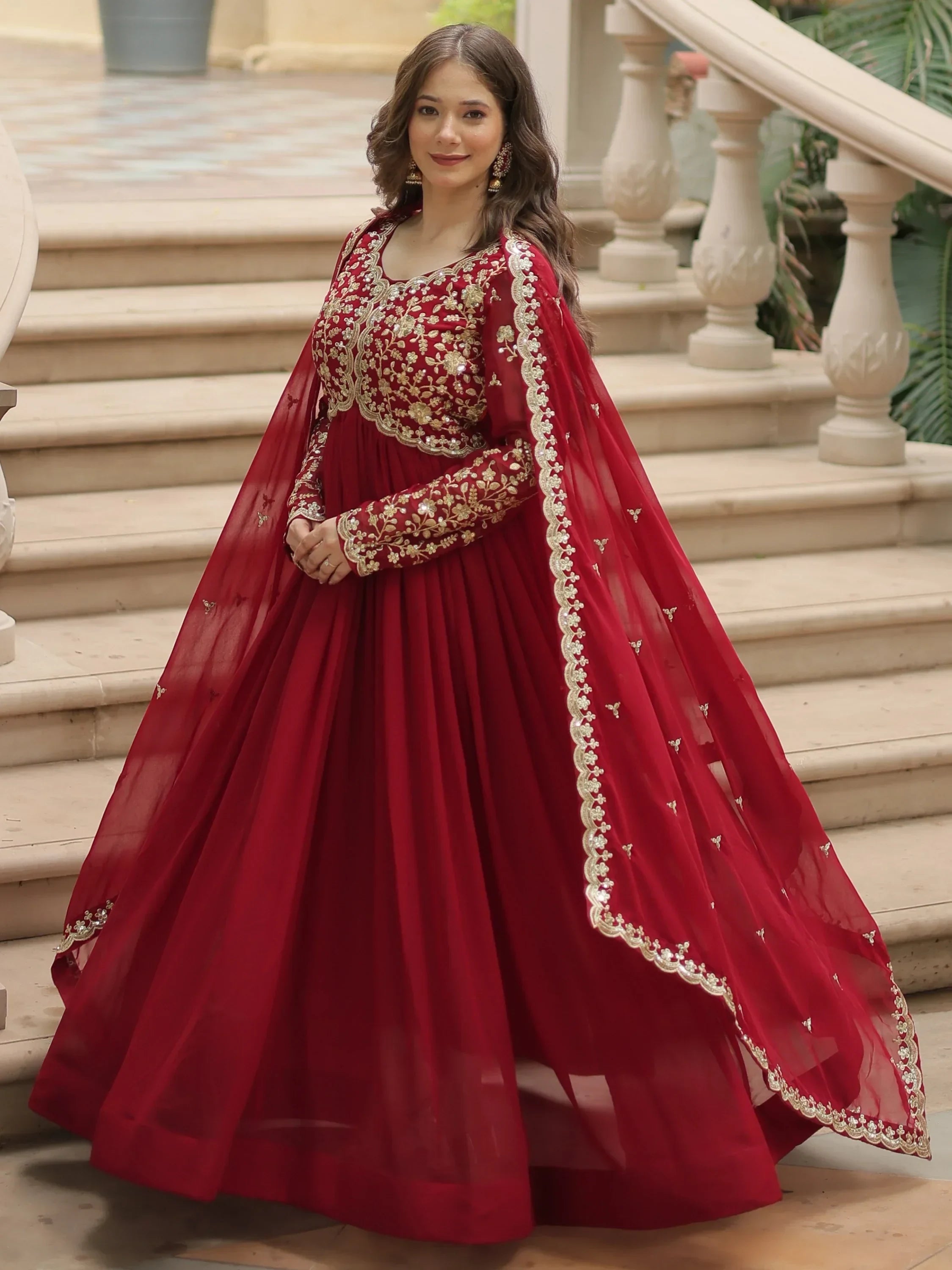 Woman in a red embroidered Indian ethnic gown with gold details, posing on stairs
