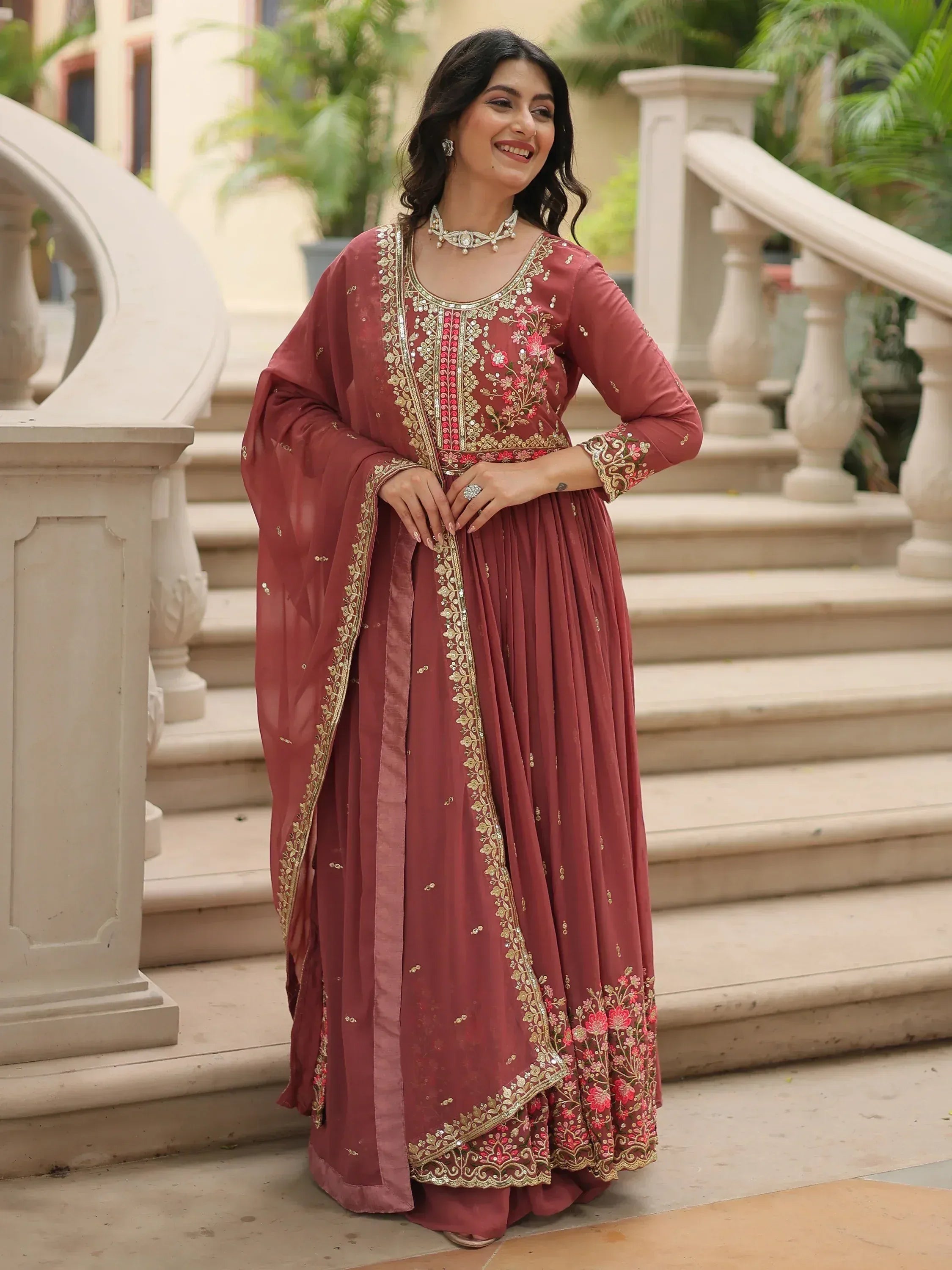Woman in maroon embroidered Indian ethnic gown with gold floral detailing, standing on stairs