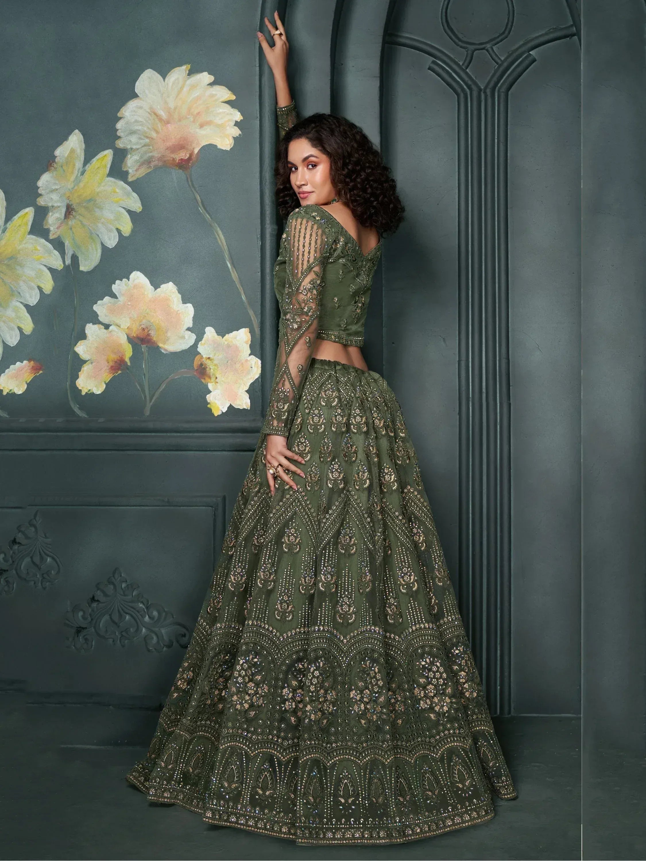 Olive green designer lehenga choli with heavy gold embroidery, sheer sleeves, and floral wall backdrop
