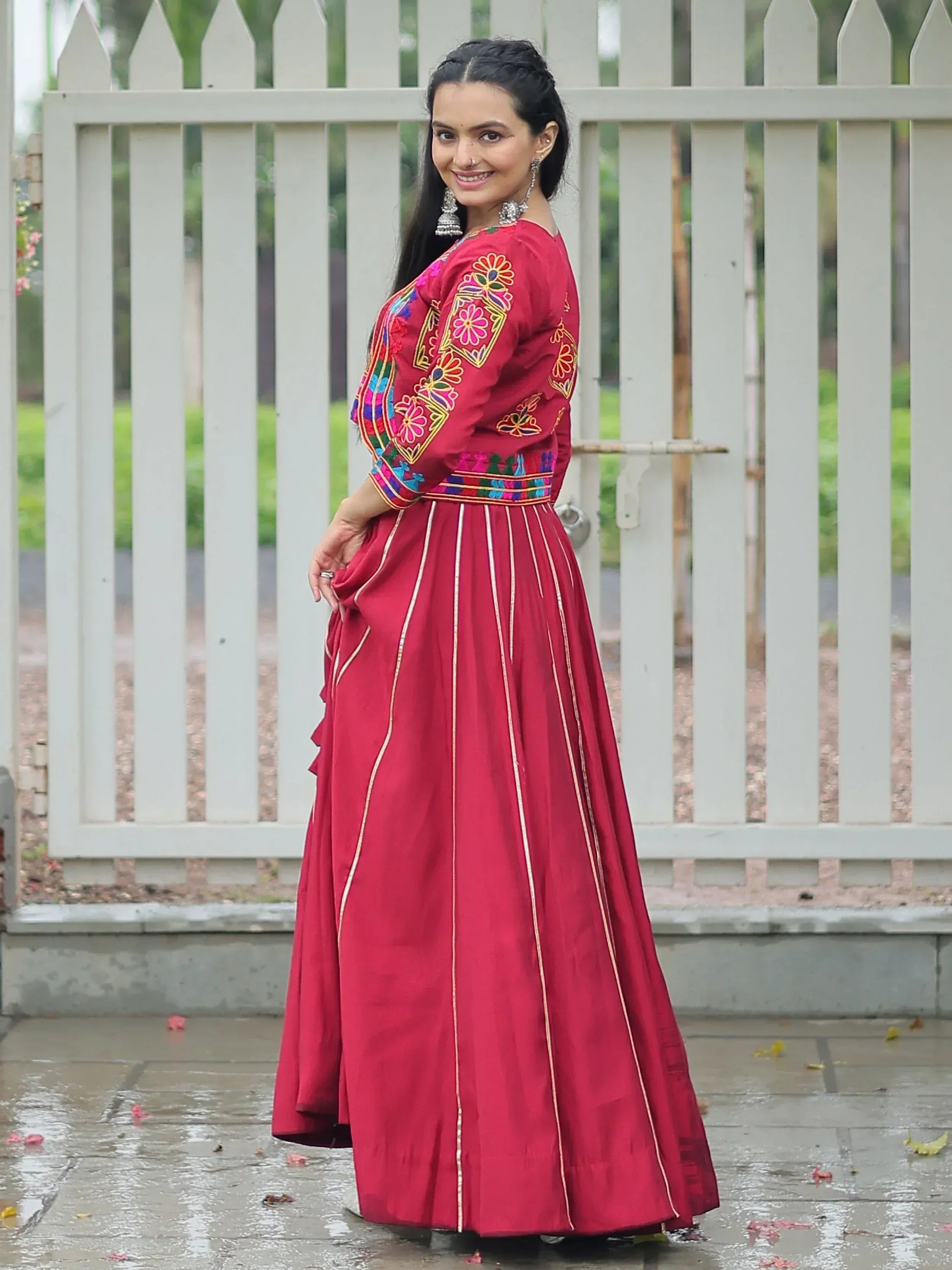Woman in maroon embroidered ethnic gown with floral motifs, gold trim, and jhumka earrings, standing by white fence.