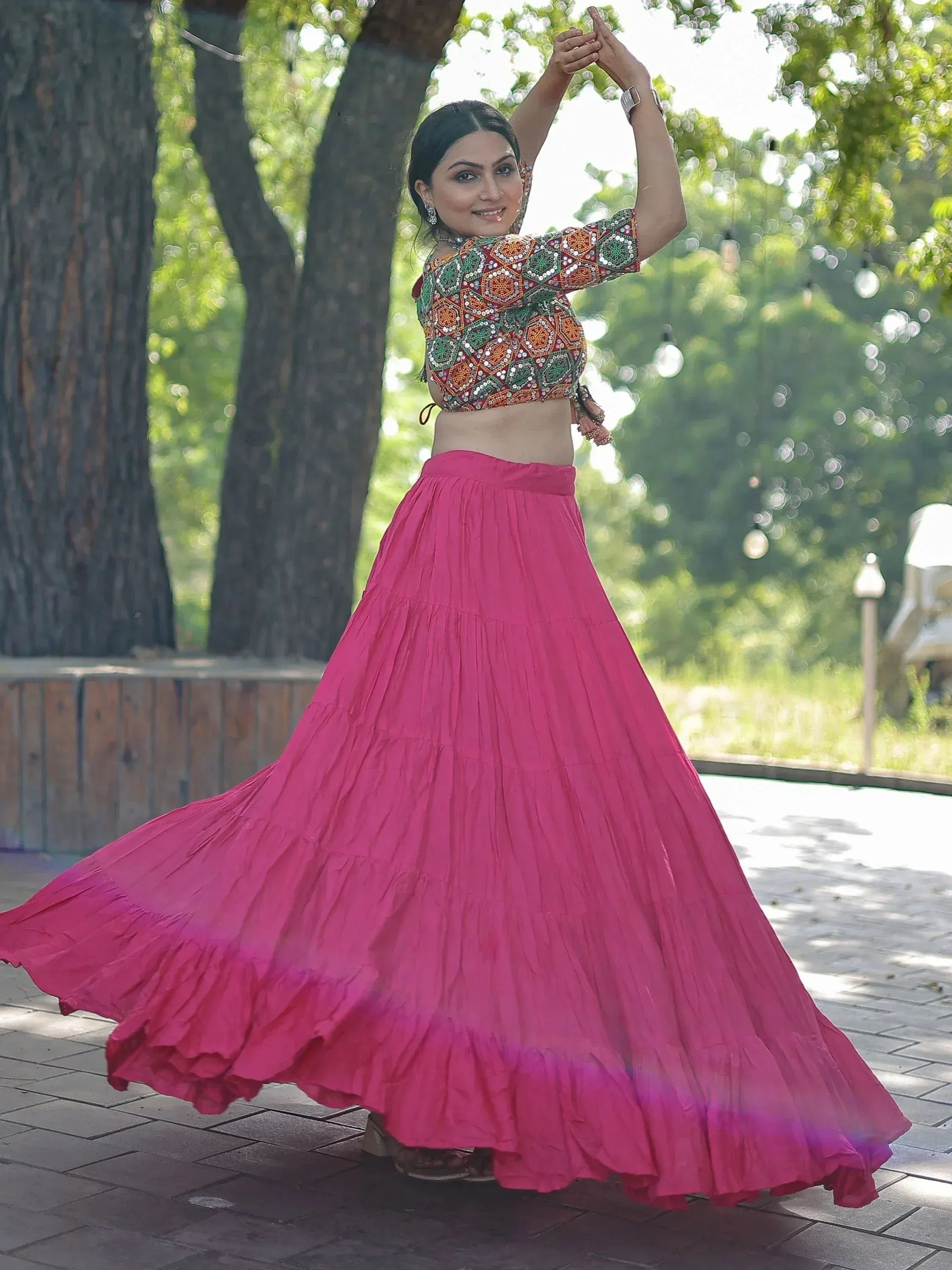 Woman in vibrant pink lehenga with embroidered crop top, dancing outdoors under trees