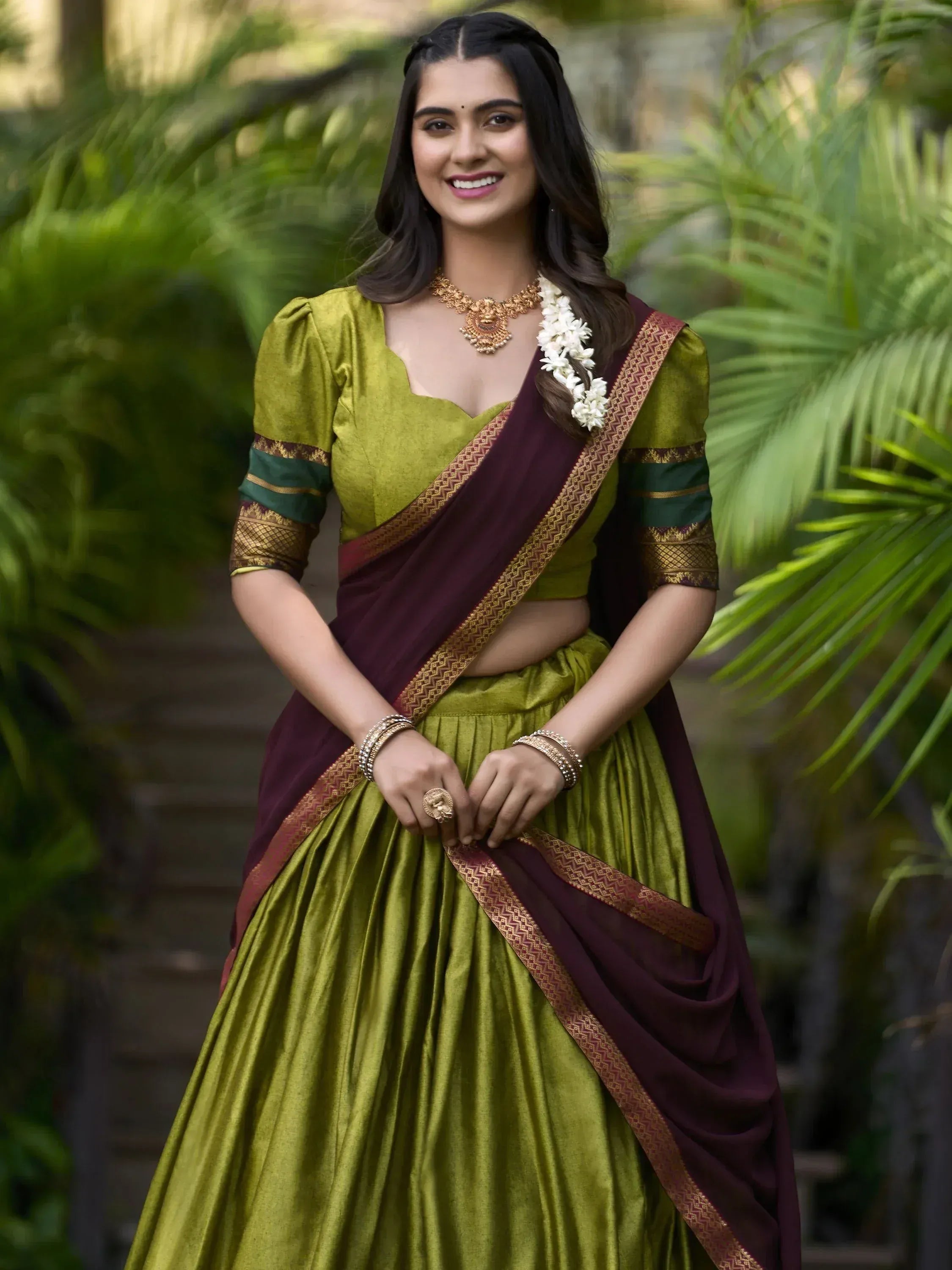 Woman in olive green and maroon lehenga choli with gold jewelry, floral hair accessory, garden backdrop