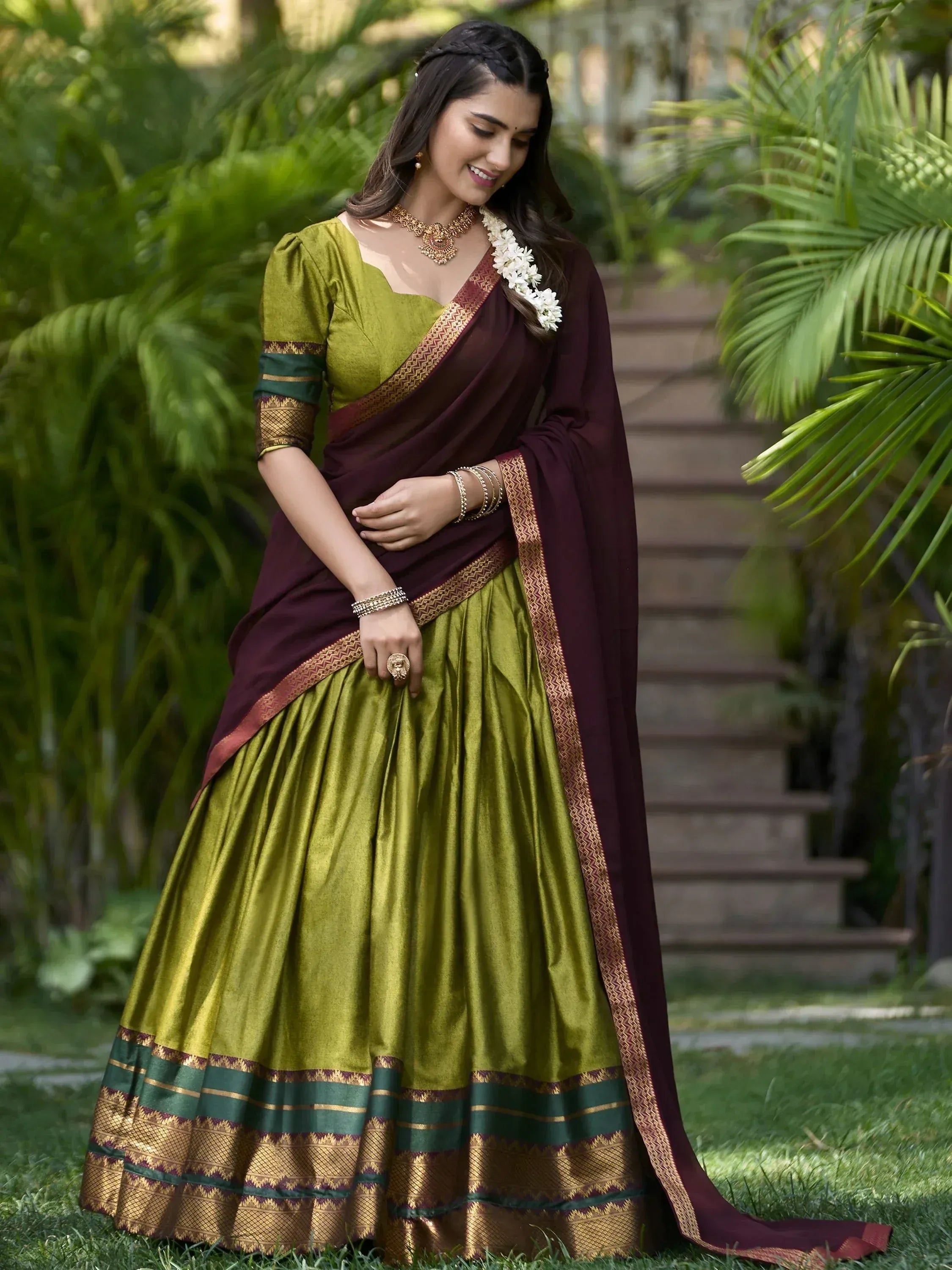 Woman in olive green and maroon ethnic saree with gold border, styled with floral garland and jewelry outdoors