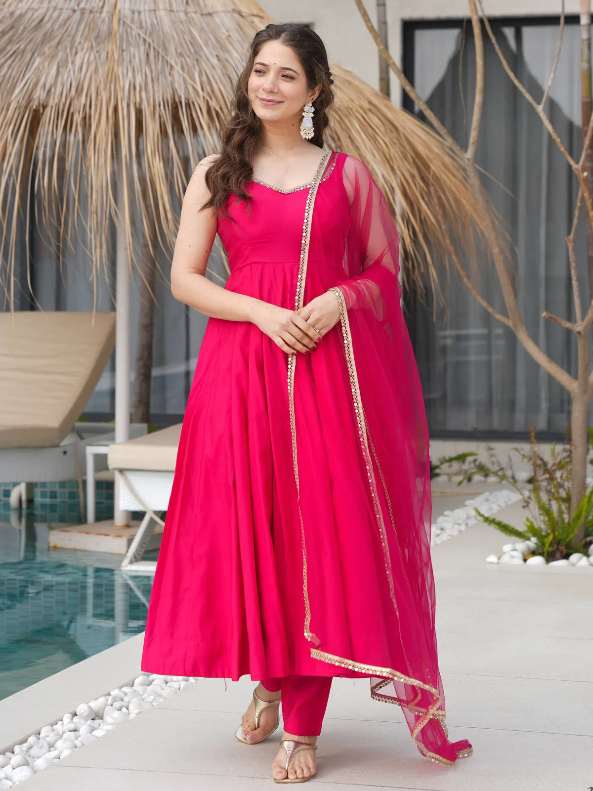 Vibrant pink ethnic gown with gold embroidery, paired with matching pants and sheer dupatta, perfect for festive occasions.