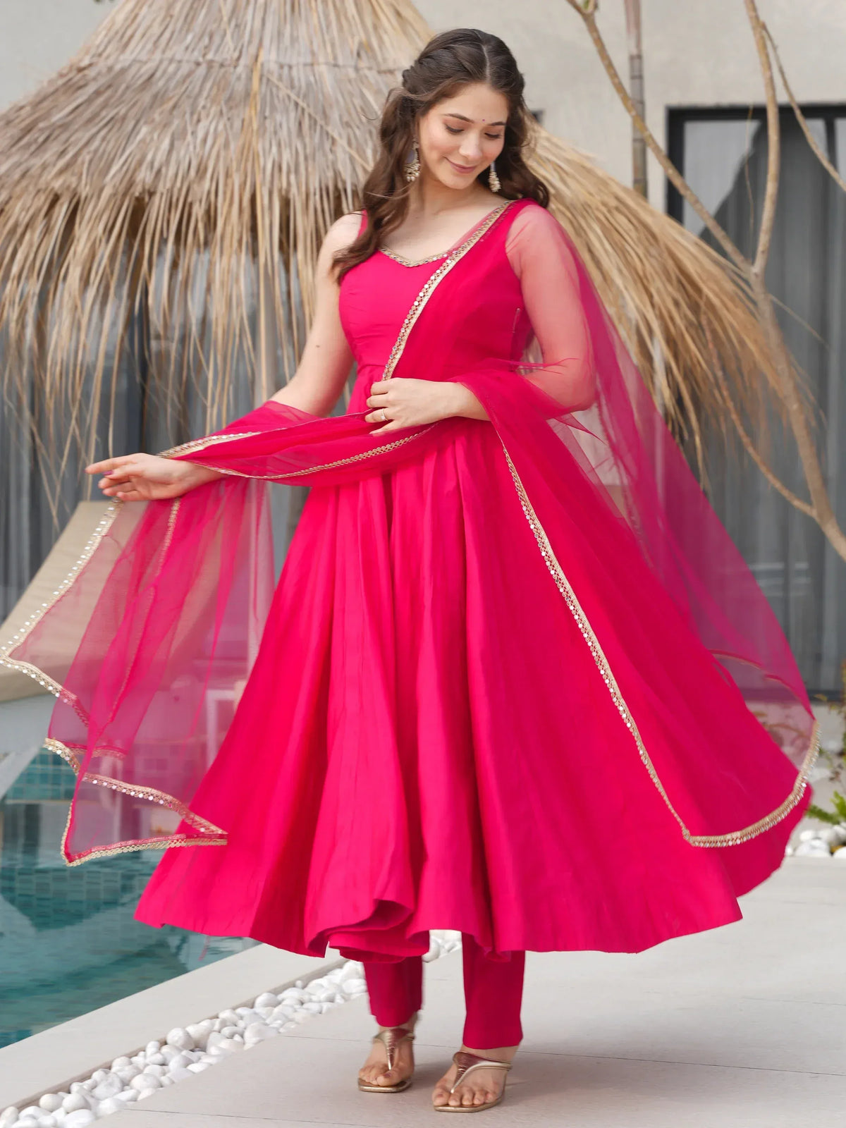 Woman in vibrant pink Indian ethnic gown with sheer dupatta, gold trim, poolside setting