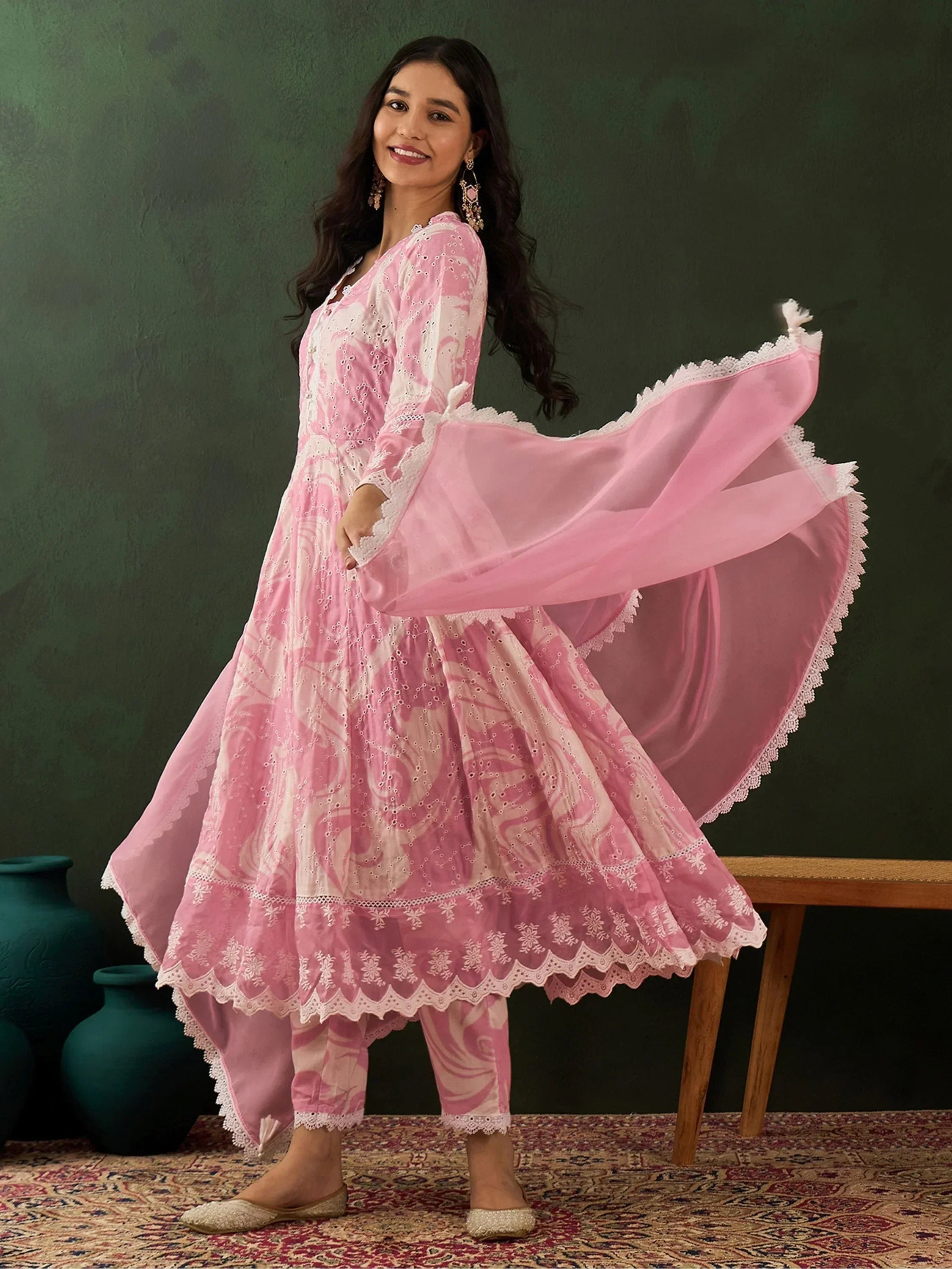 Woman in a pink embroidered Anarkali suit with dupatta, standing on a patterned rug.