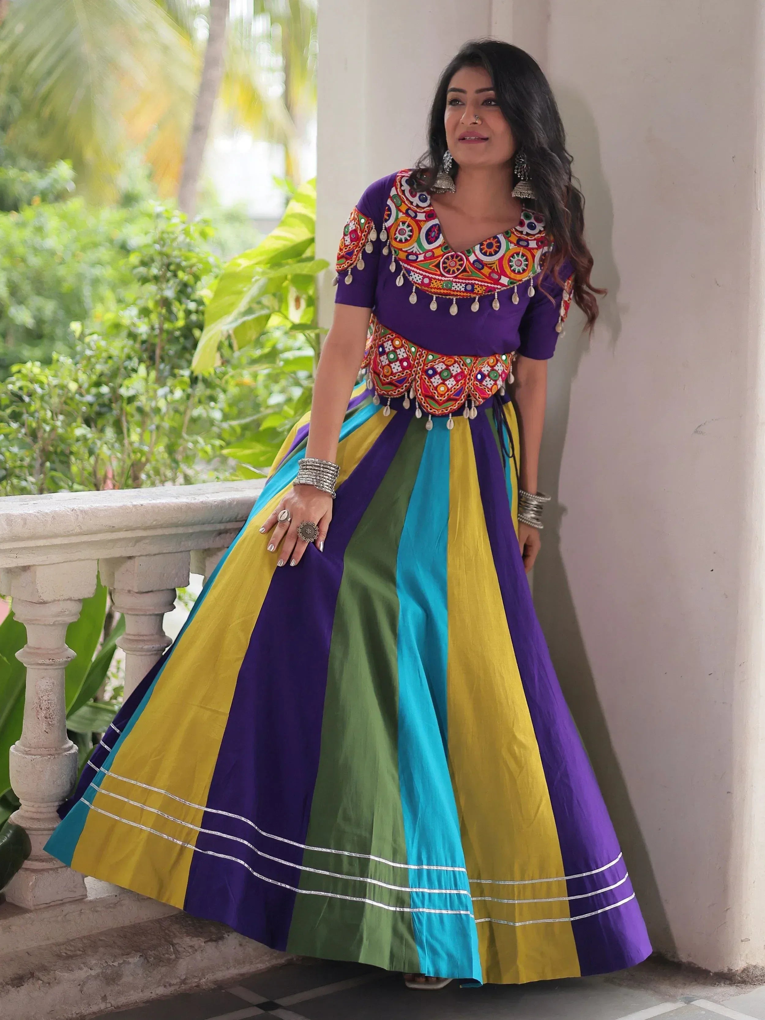Woman in vibrant purple, yellow, green, and blue ethnic lehenga with mirror work and embroidery, perfect for Indian festivals.