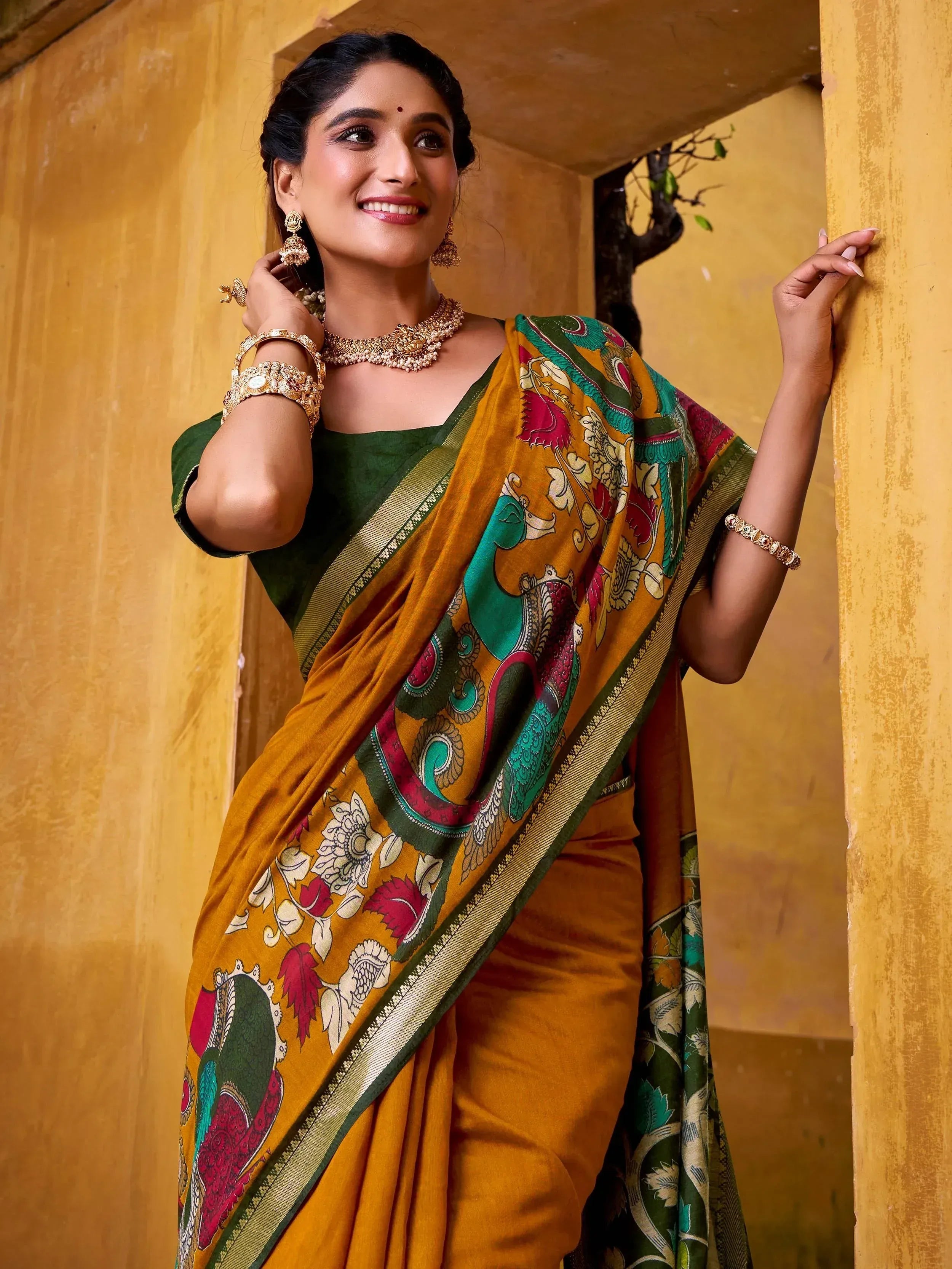 Woman in vibrant printed saree with green blouse, gold jewelry, against yellow wall