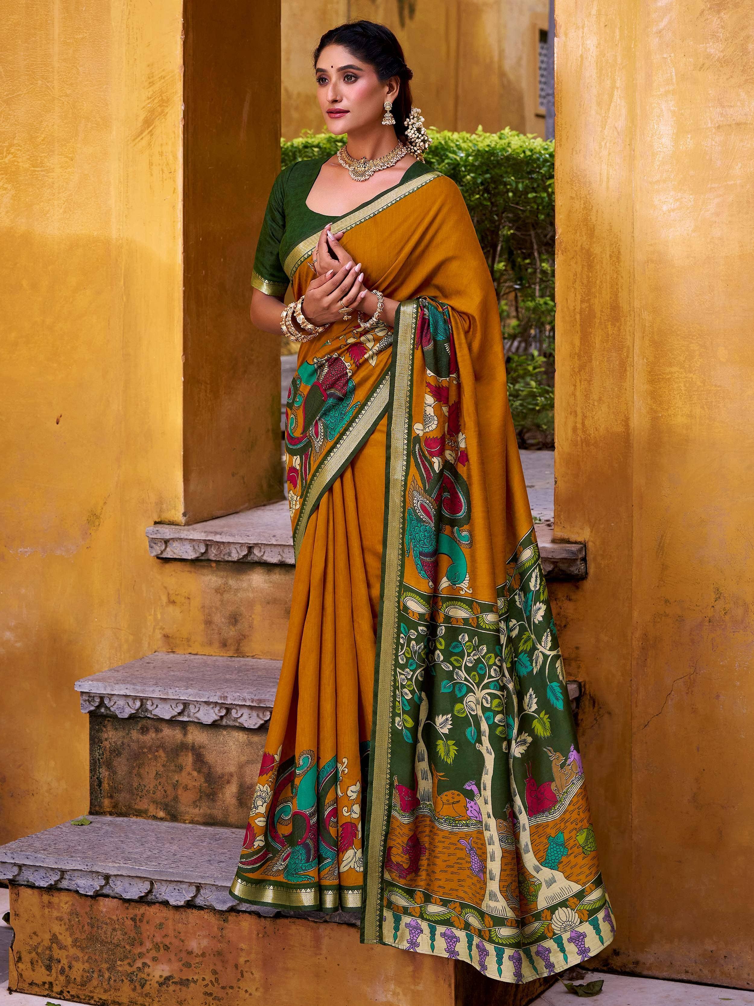 Woman in mustard yellow saree with green blouse and vibrant printed border, styled with gold jewelry.