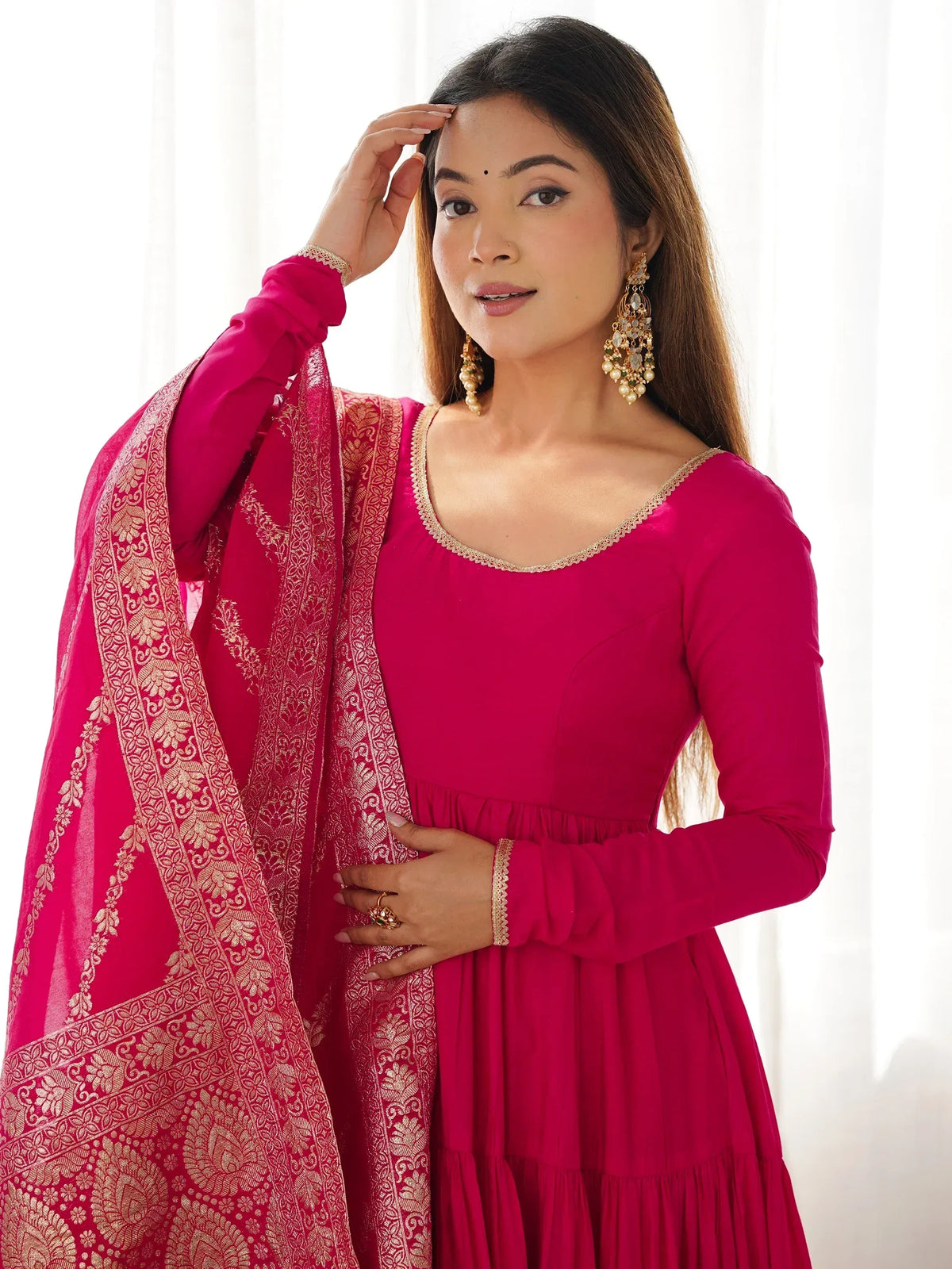 Woman in vibrant pink ethnic gown with gold embroidery, paired with matching dupatta and traditional jewelry