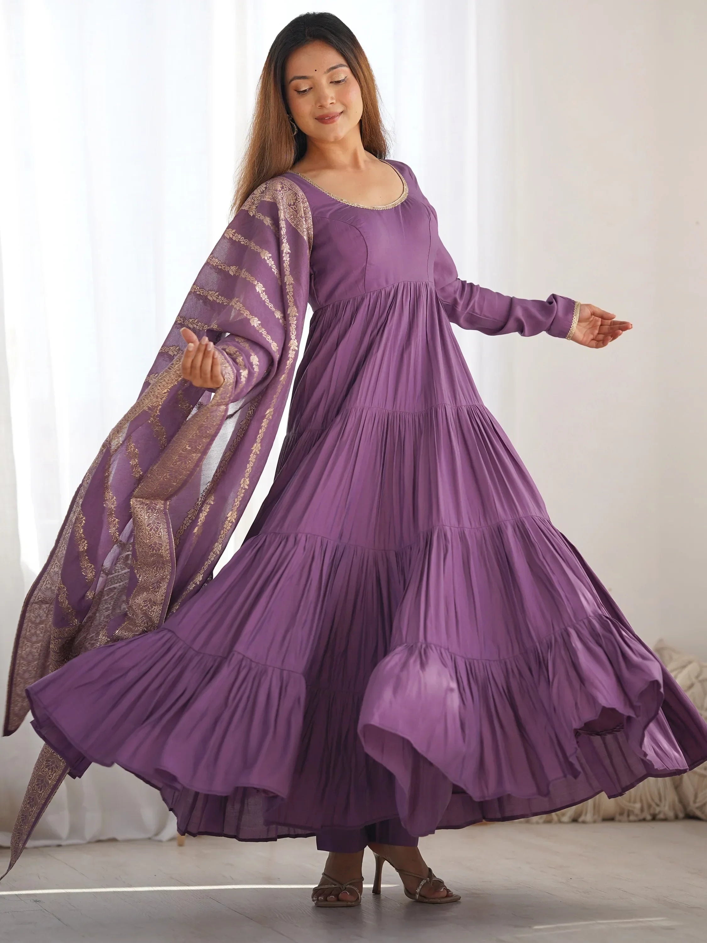 Woman wearing a flowy purple Anarkali dress with gold embroidered dupatta indoors