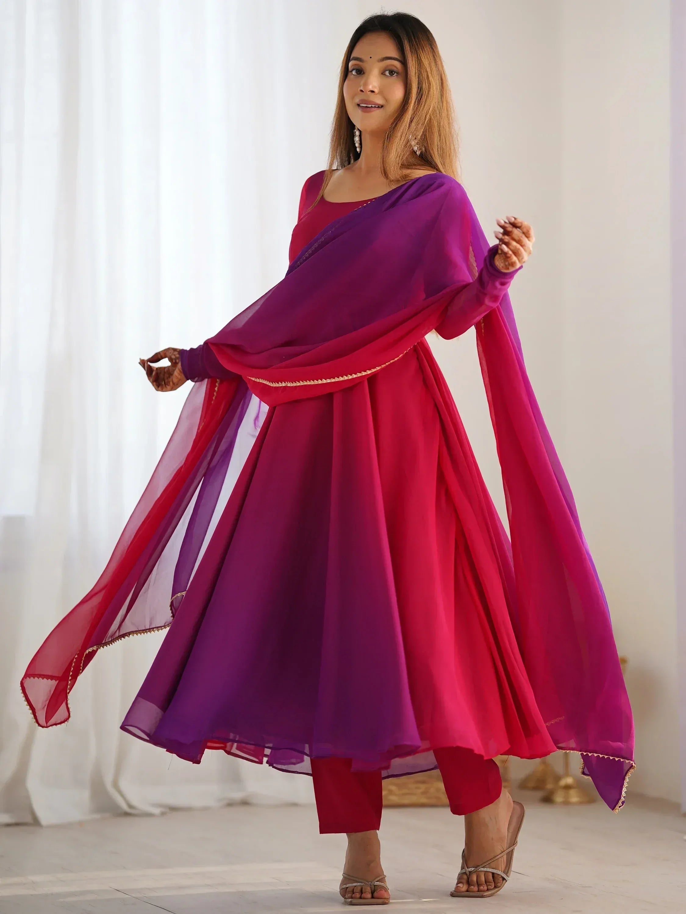 Woman in vibrant pink and purple Indian ethnic anarkali suit with sheer dupatta, perfect for festive occasions.