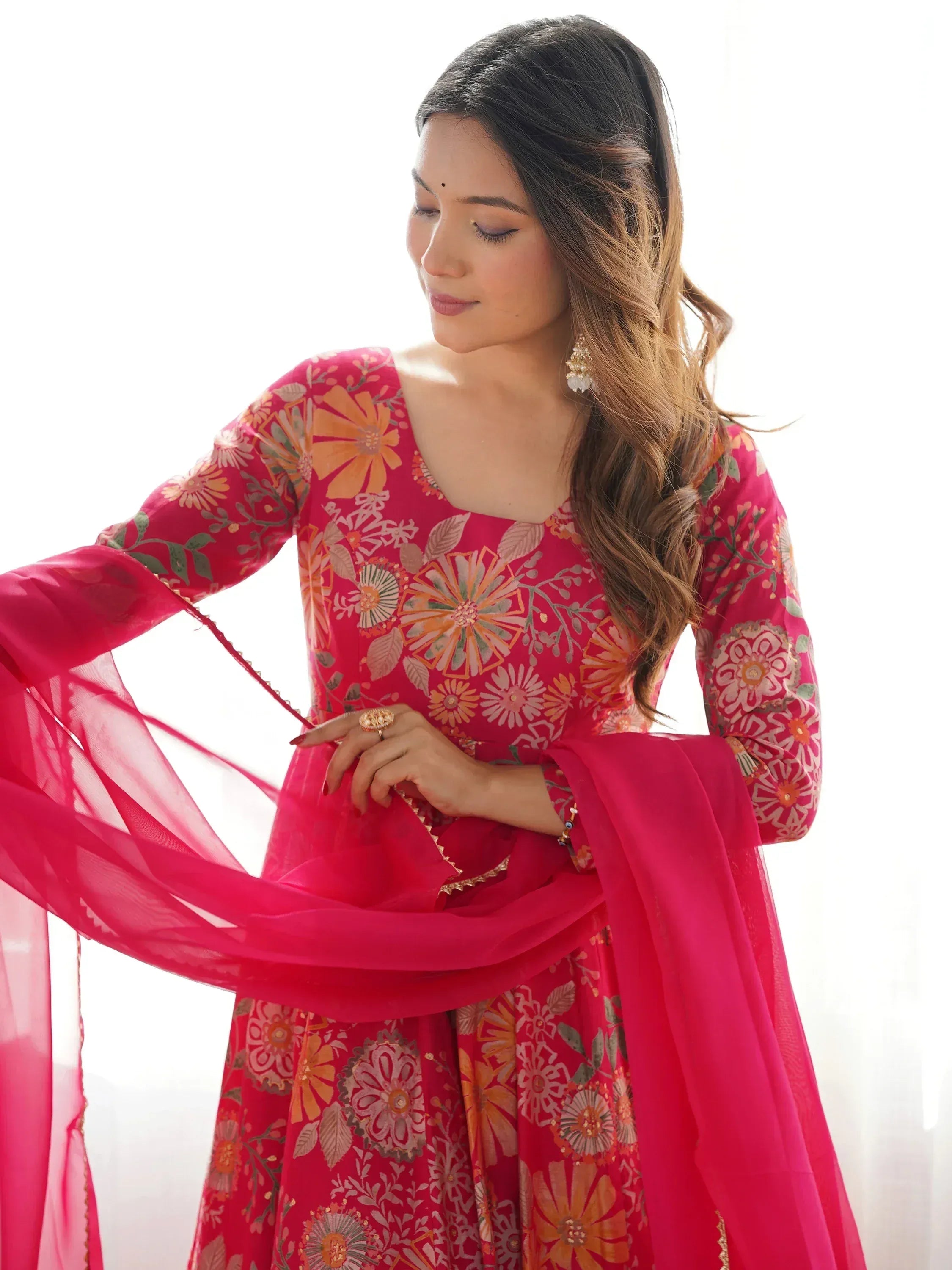 Woman in vibrant pink floral print Indian ethnic gown with sheer dupatta, perfect for festive occasions.
