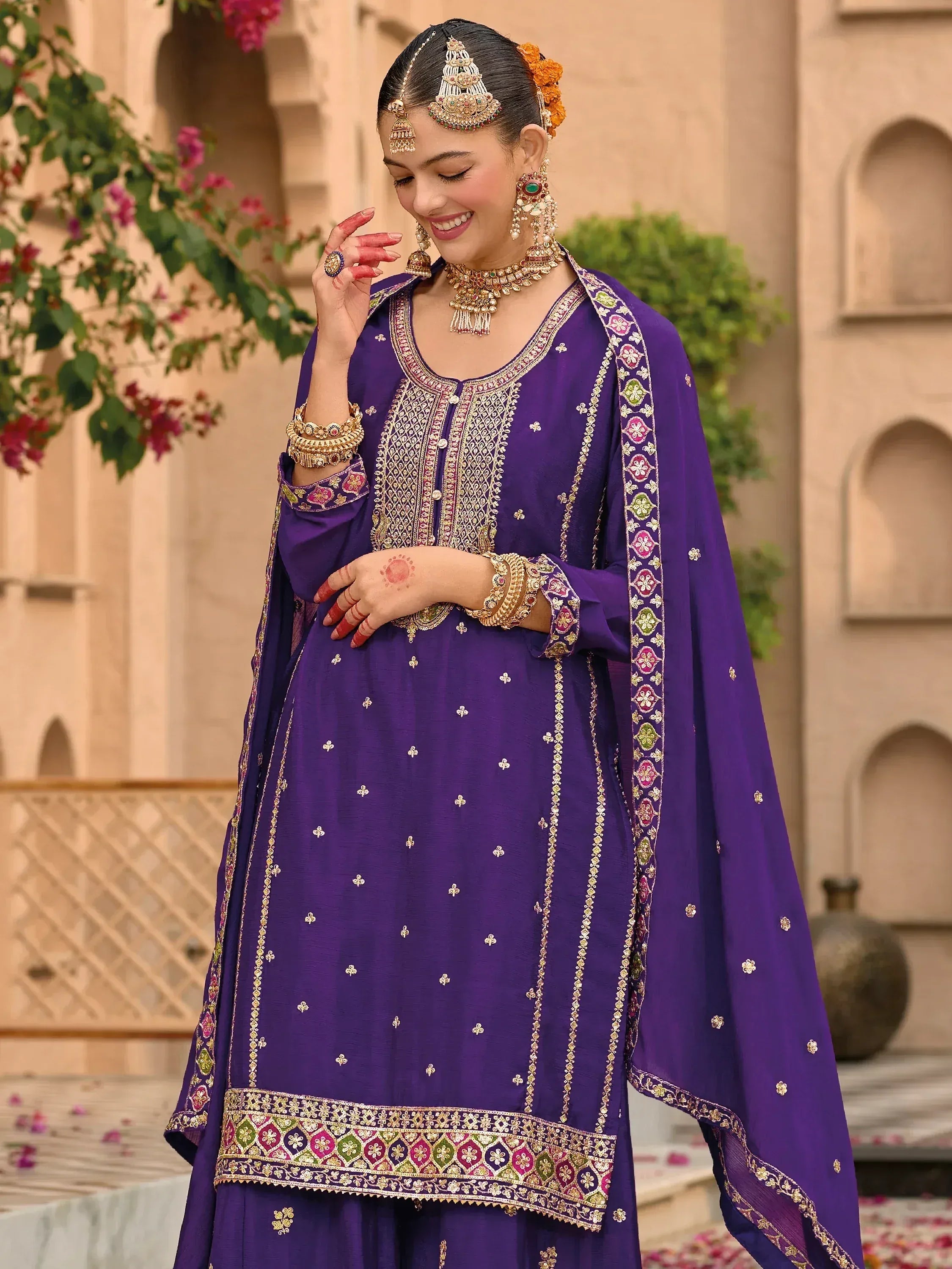 Purple embroidered Indian salwar kameez with gold detailing, traditional jewelry, and floral dupatta for festive occasions