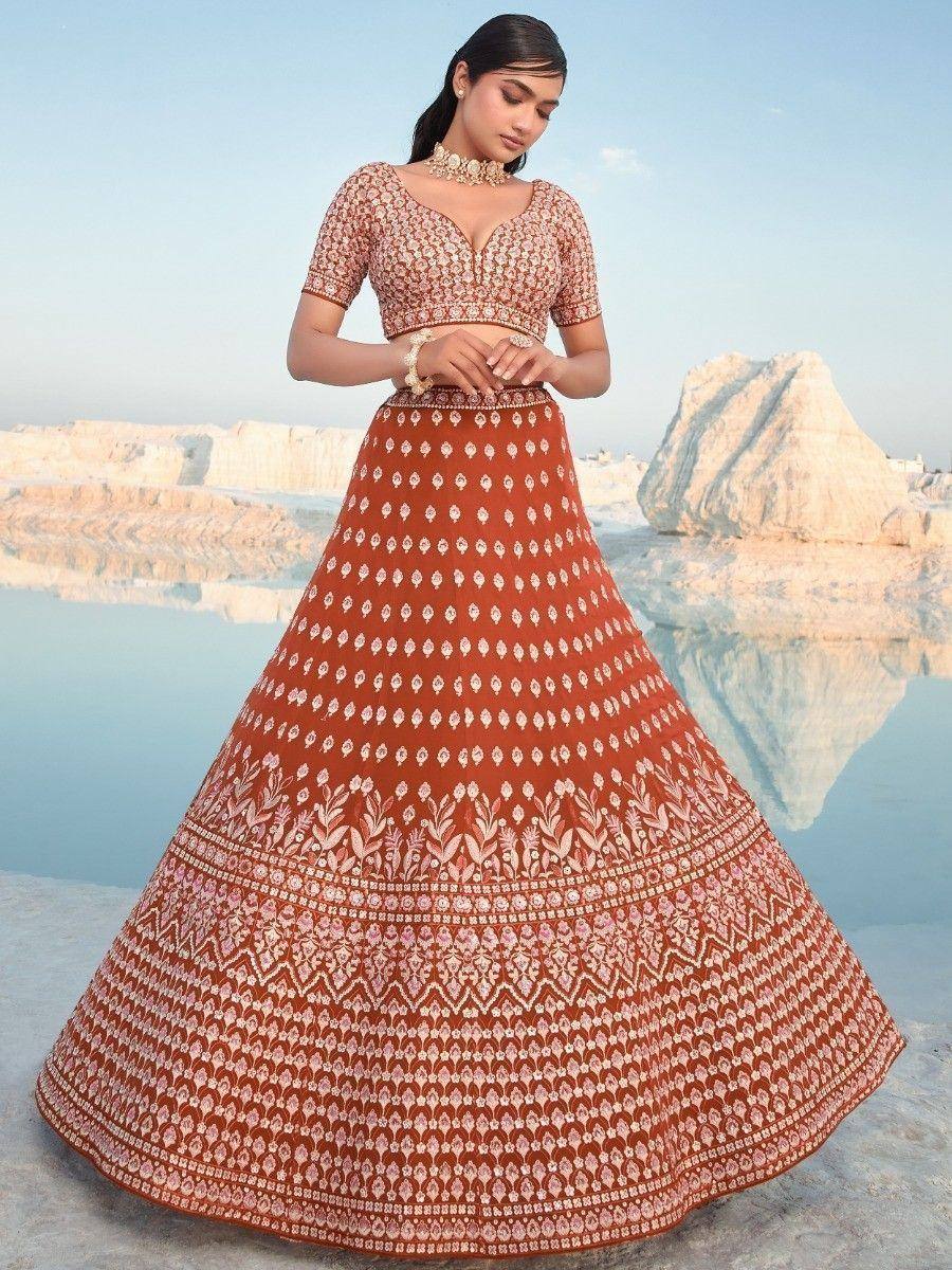 Woman in rust-colored printed lehenga choli with floral motifs, standing by water against white rock formations