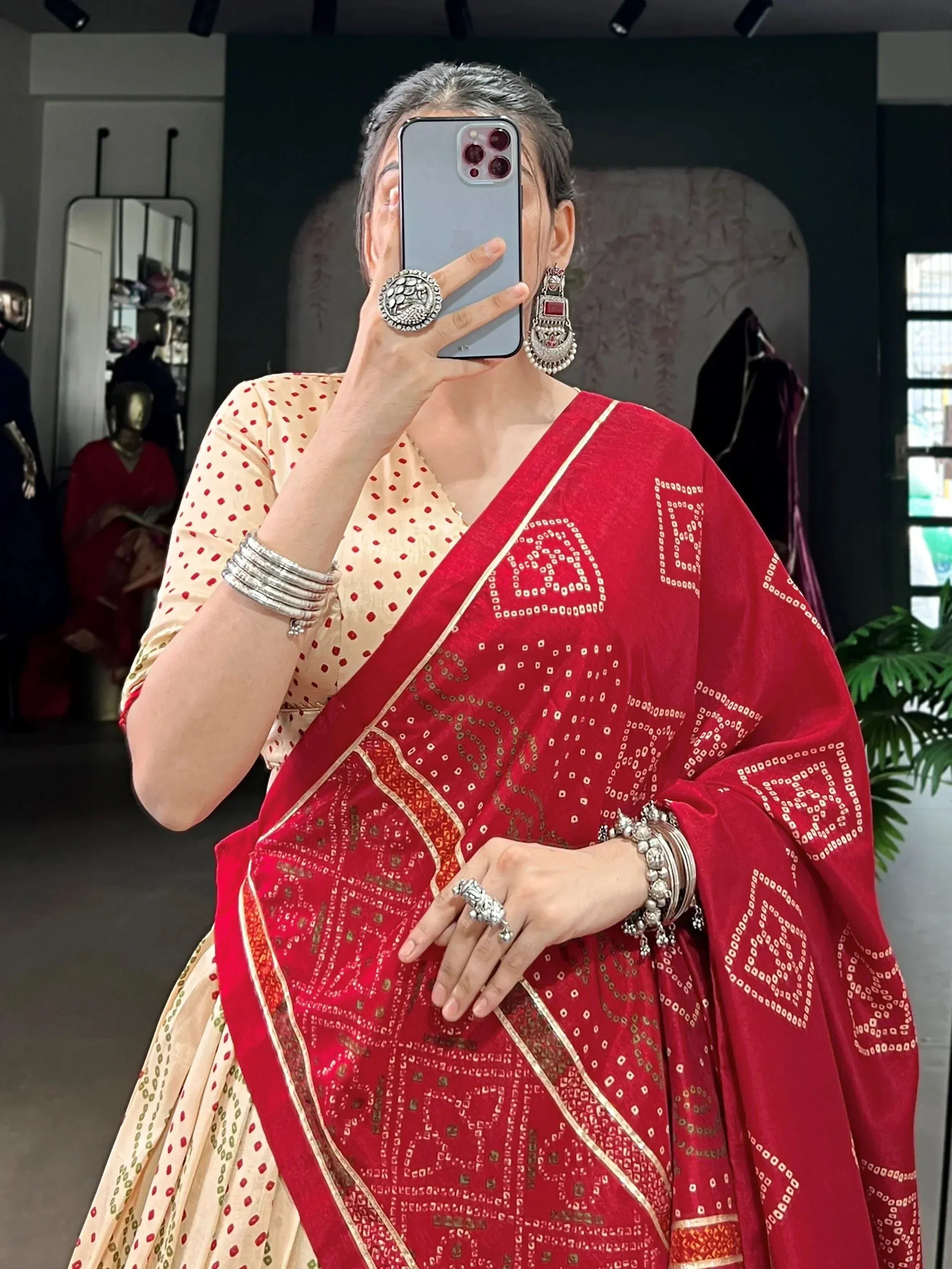 Woman in red printed saree with gold border, ethnic jewelry, taking mirror selfie in boutique