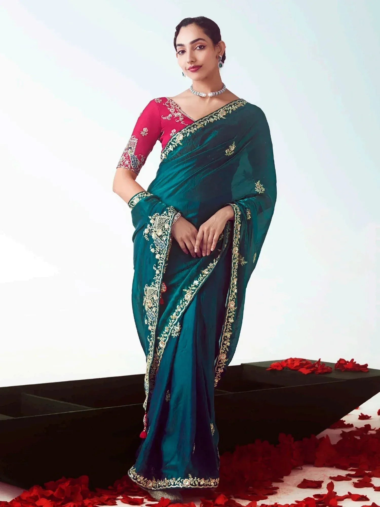 Elegant teal saree with gold embroidery and pink blouse, styled with jewelry and rose petals for festive occasions.