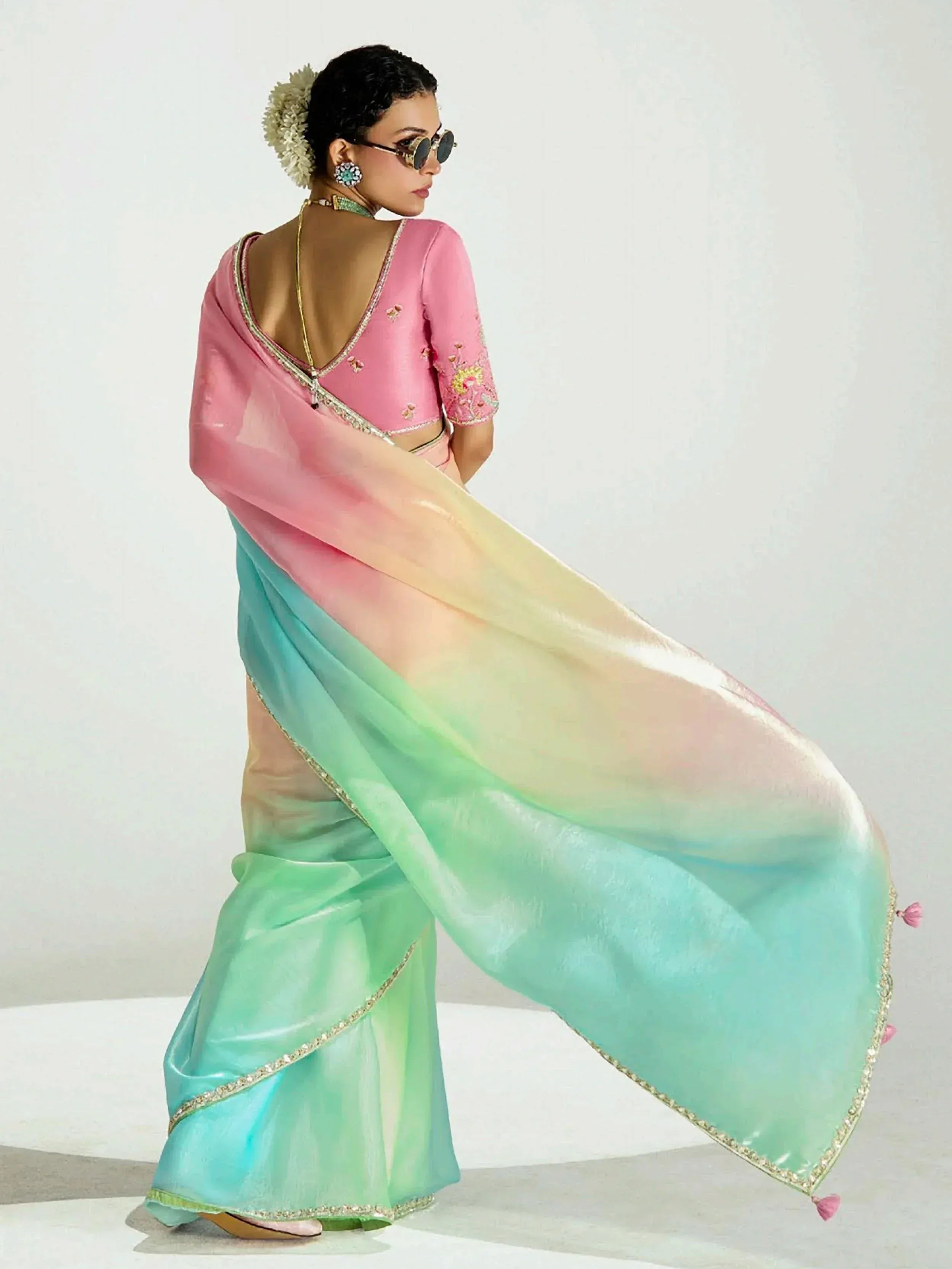 Woman in pastel ombre saree with pink blouse, floral embroidery, and gold trim, styled with sunglasses and flowers.