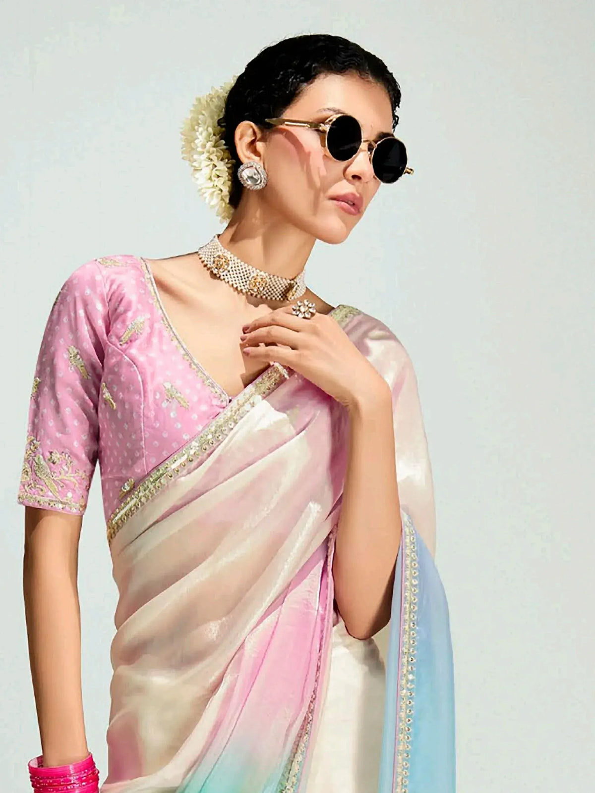 Woman in elegant pink and ombre saree with gold embroidery, styled with sunglasses and floral hair accessory.