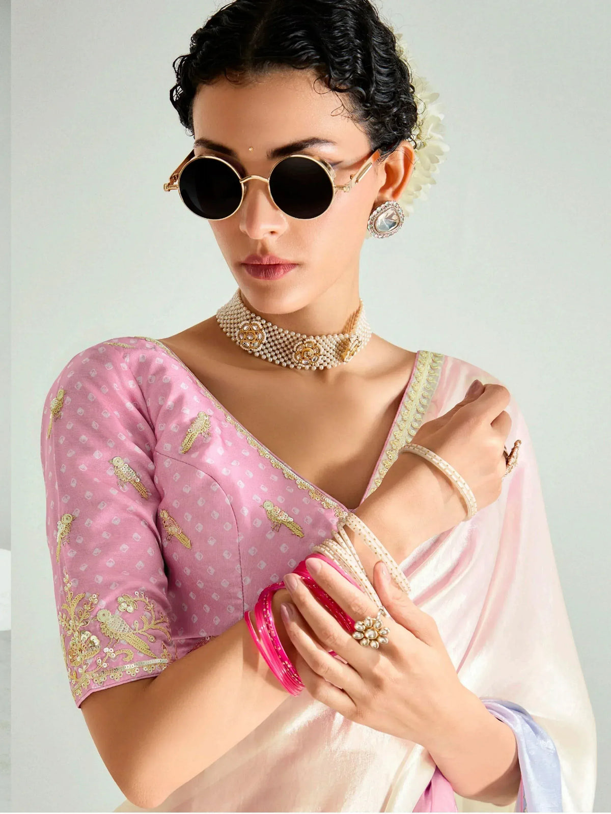 Woman in pink embroidered saree with gold bird motifs, pearl jewelry, and round sunglasses