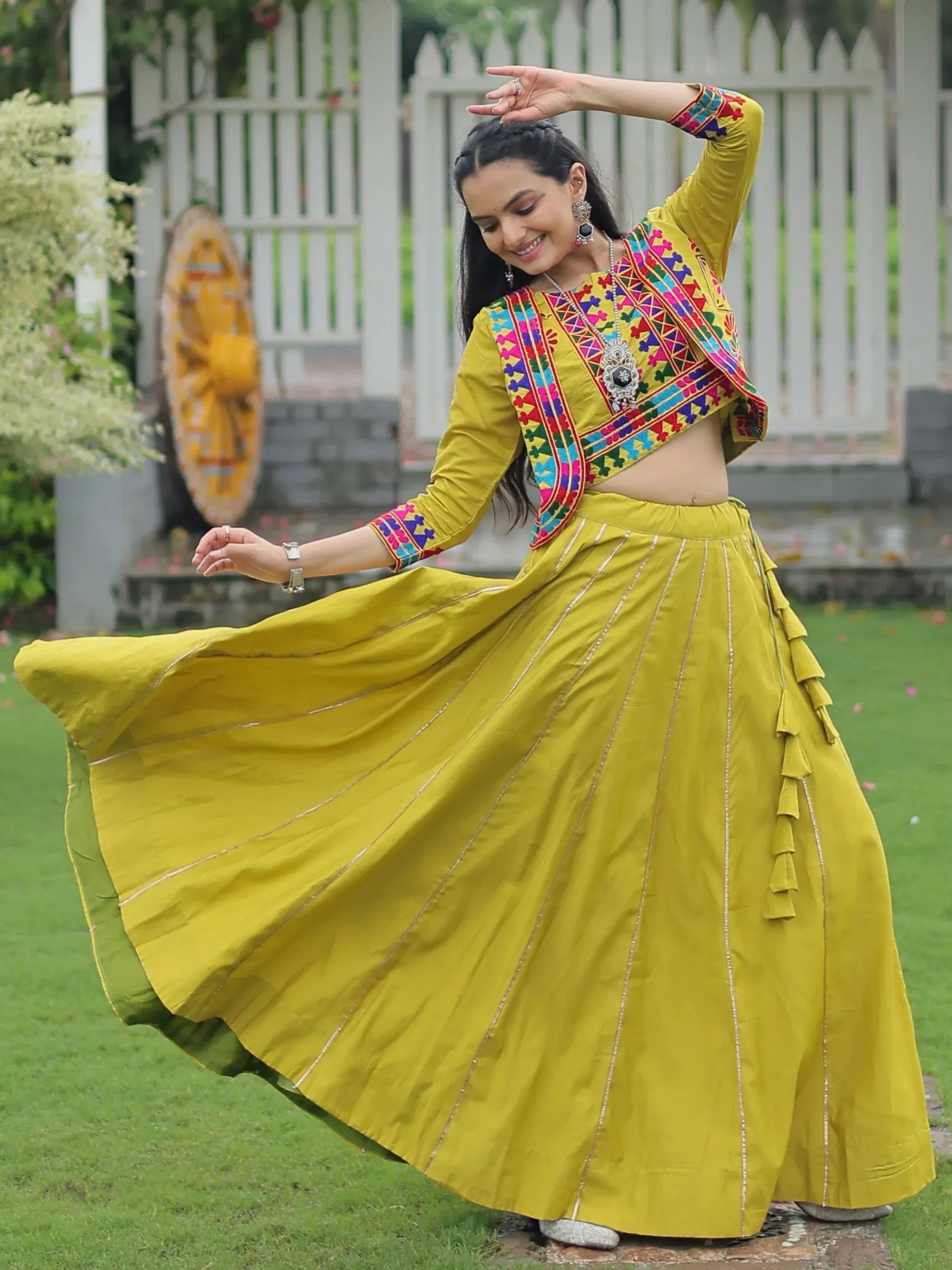 Woman in vibrant yellow ethnic wear with colorful embroidery, twirling outdoors in a garden setting.