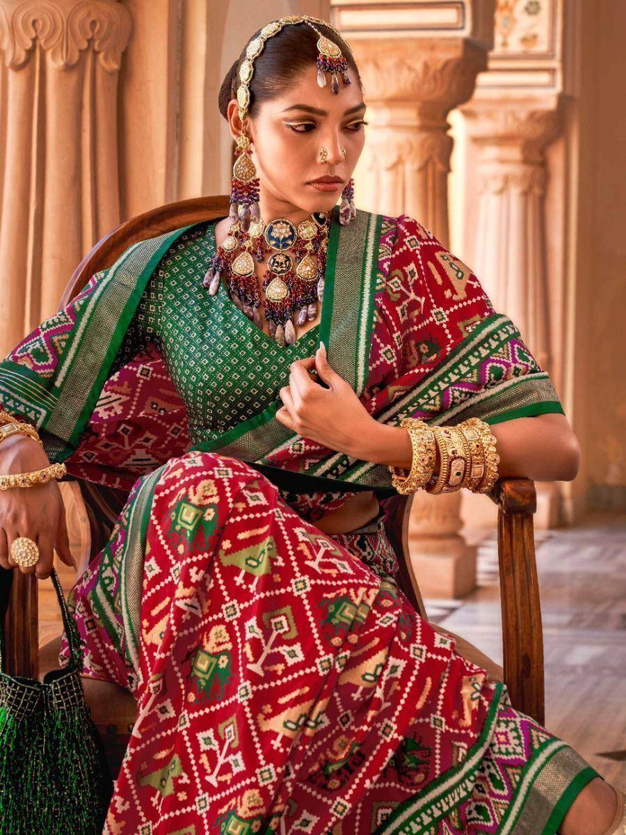Woman in red and green printed ethnic saree with traditional jewelry, seated in palace setting