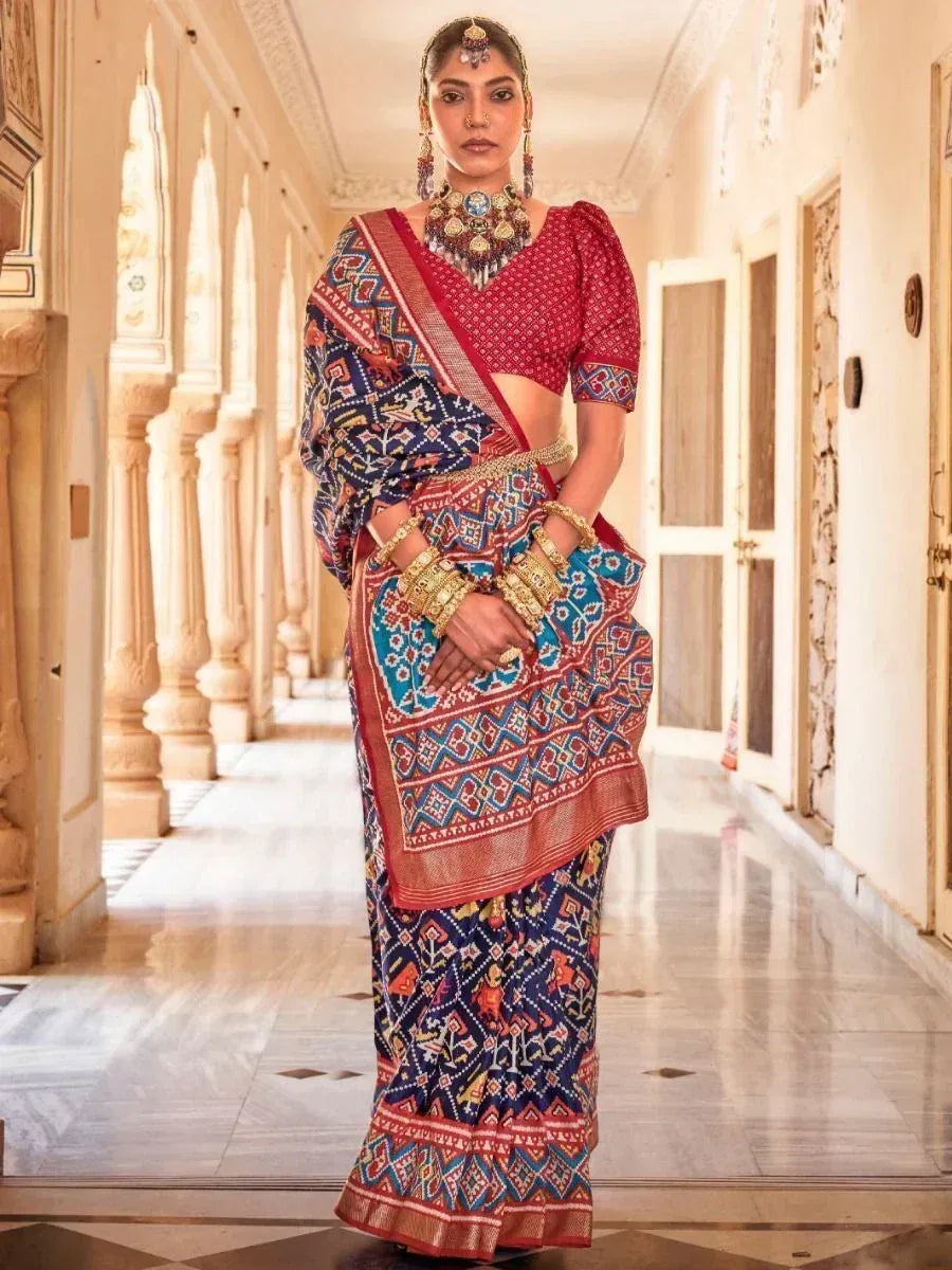 Woman in vibrant red and blue printed saree with gold jewelry, standing in ornate hallway