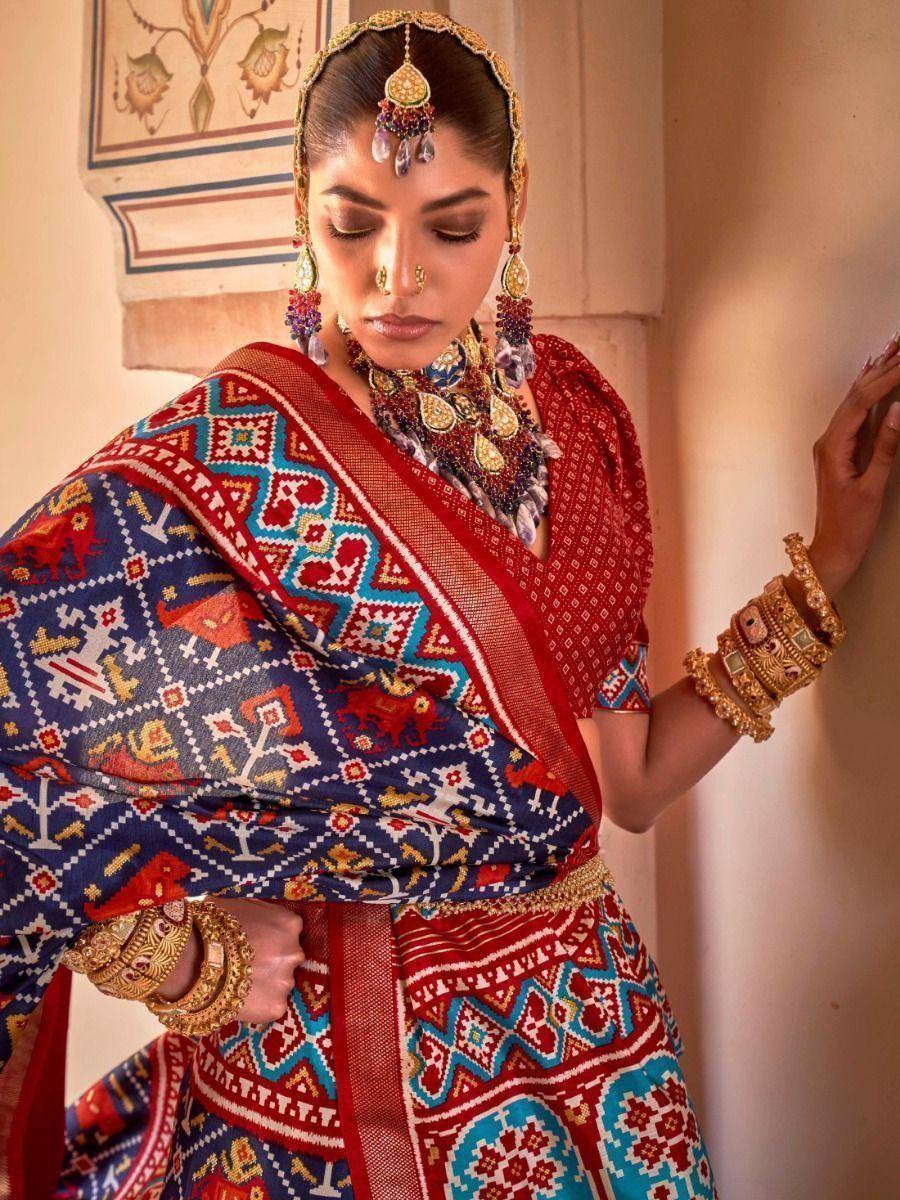 Woman in vibrant red and blue ethnic saree with intricate patterns and traditional gold jewelry
