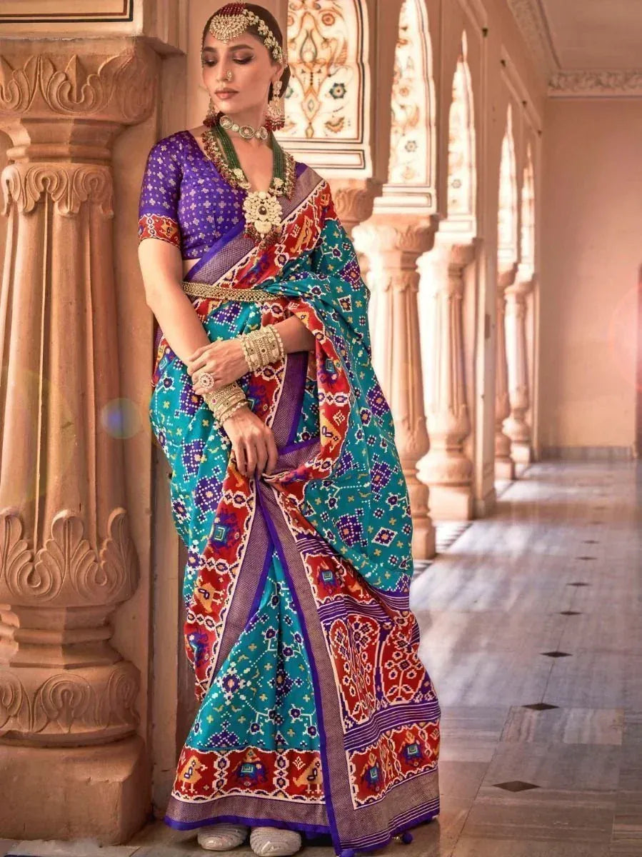 Woman in colorful Indian saree with intricate patterns, traditional jewelry, and purple blouse, posing in ornate hallway.