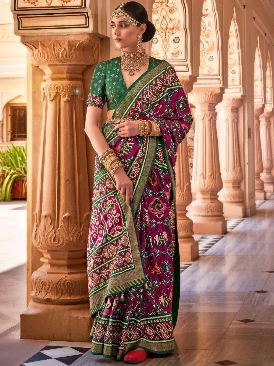 Woman in vibrant purple and green printed saree with gold jewelry, standing in ornate Indian palace hallway