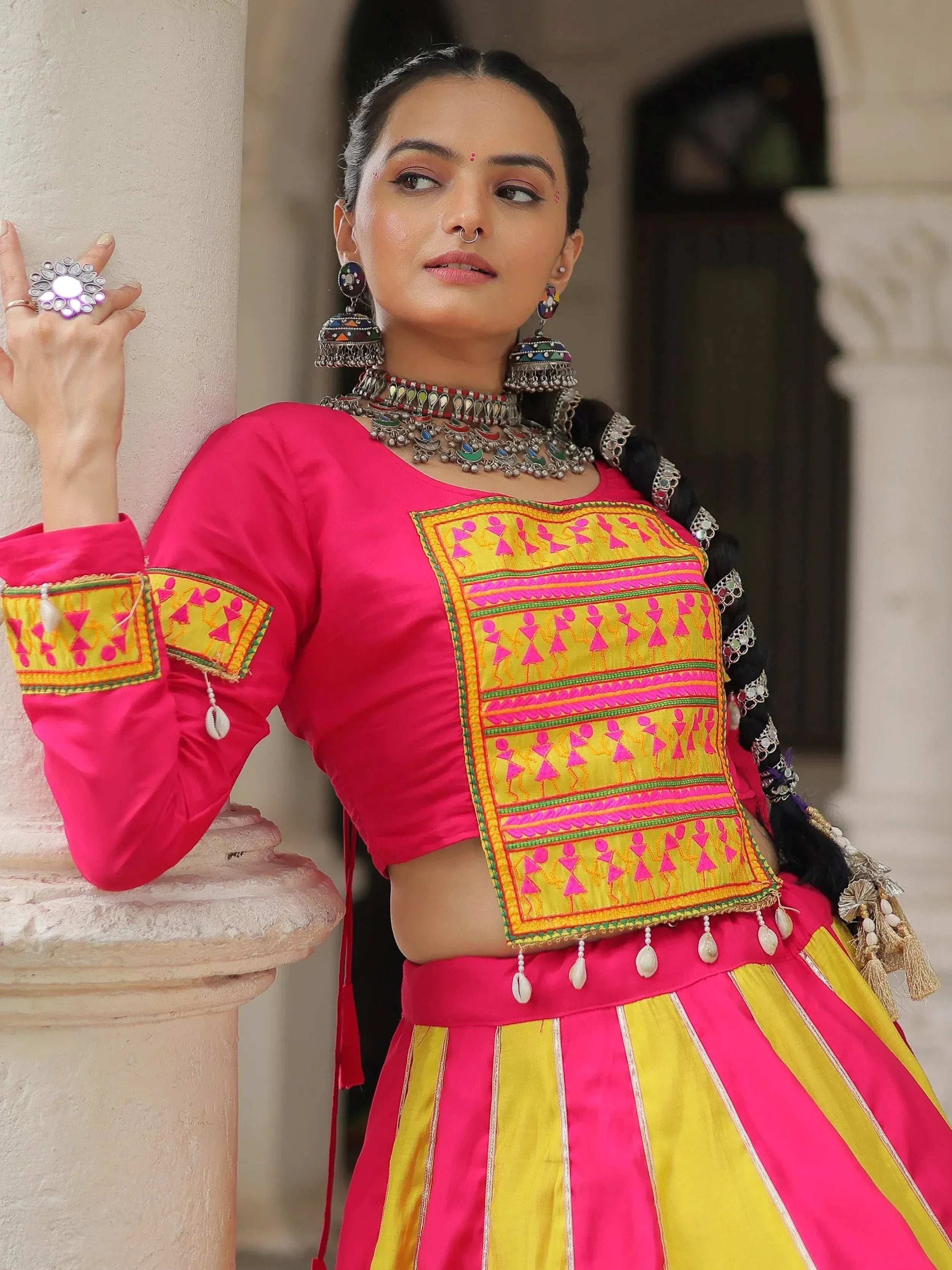Vibrant pink and yellow ethnic lehenga choli with mirror work, traditional jewelry, and embroidered details