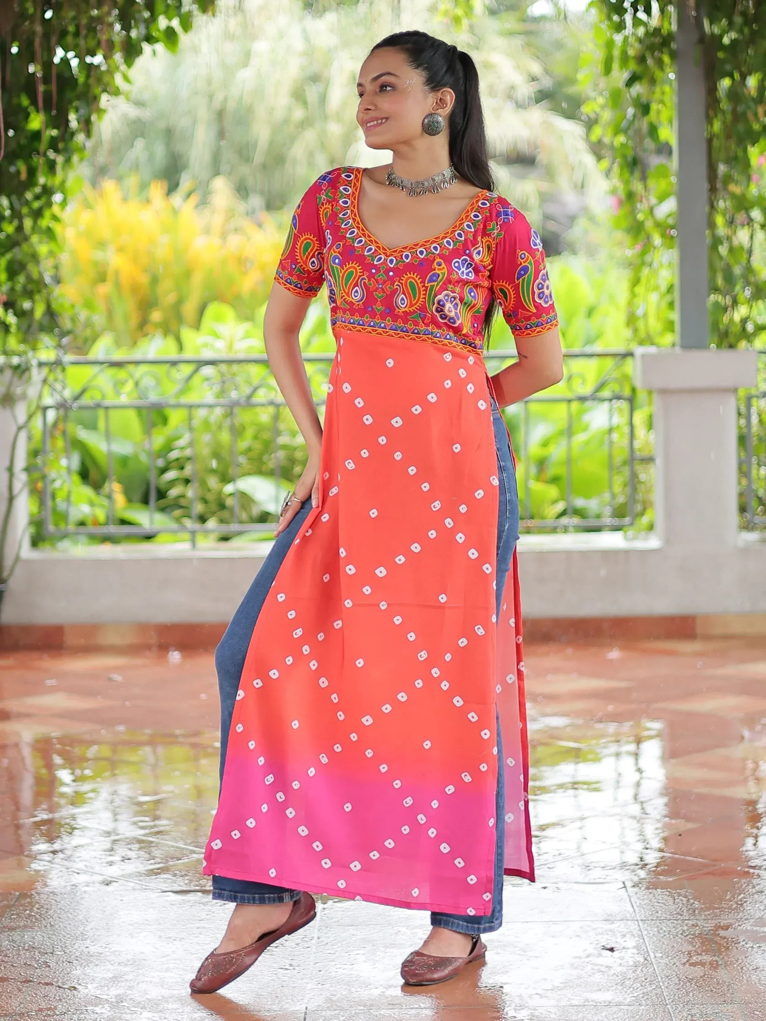 Woman in vibrant Indian ethnic kurta with paisley print, ombre orange-pink skirt, and denim pants, styled with jhumka earrings.