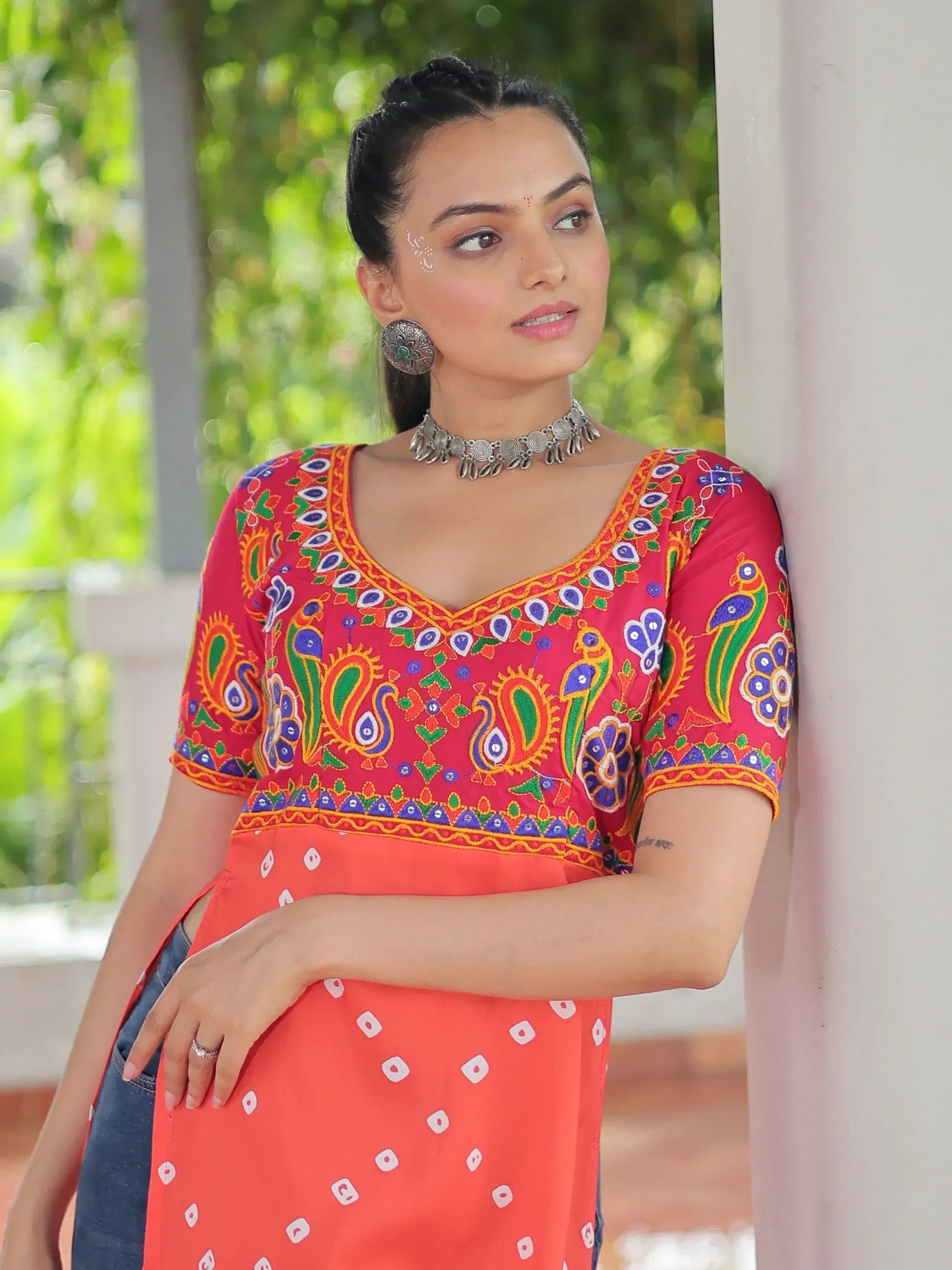 Woman in vibrant red embroidered kurta with peacock motifs, paired with orange printed skirt and silver jewelry