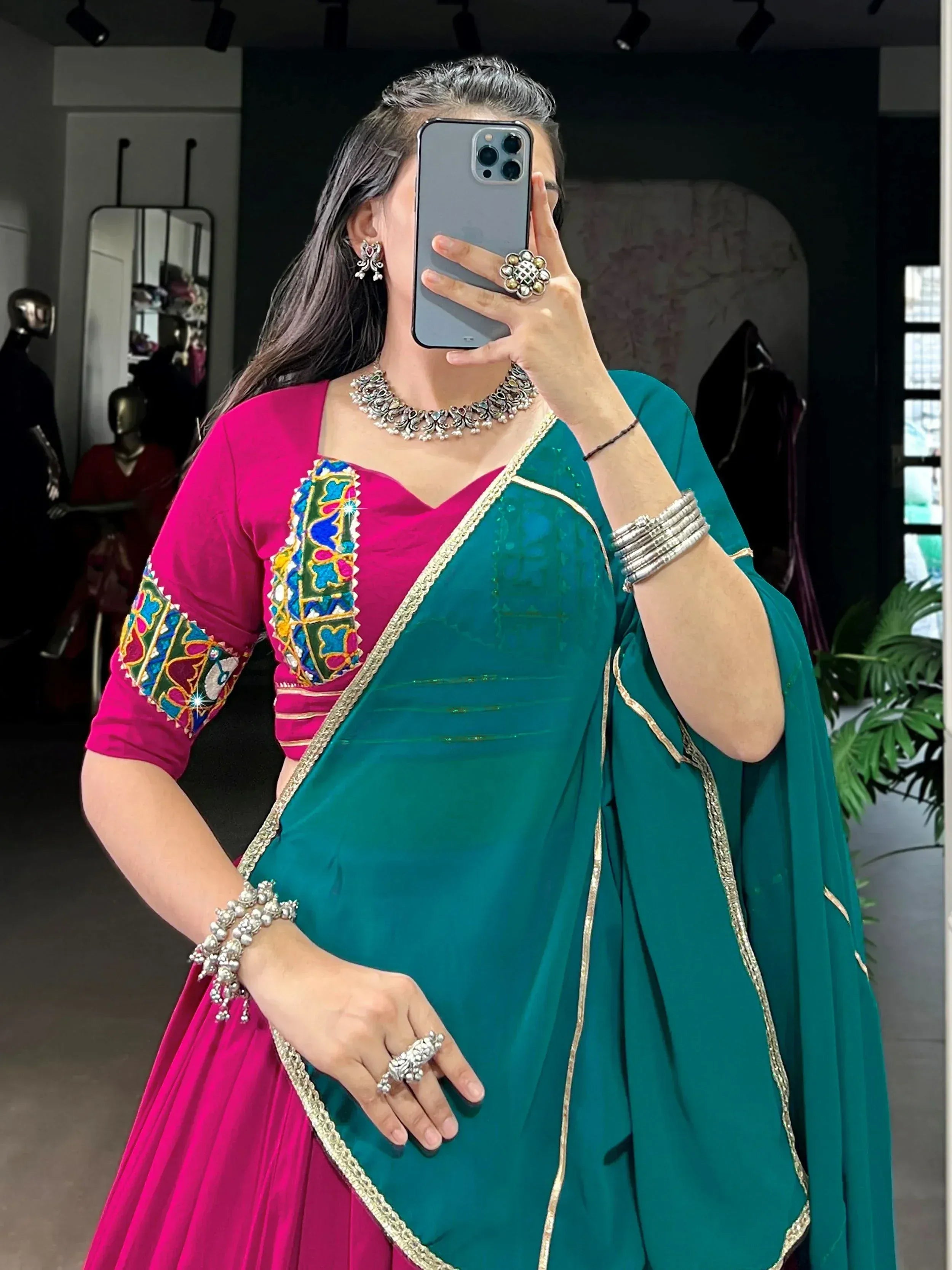 Woman in vibrant pink and teal Indian lehenga choli with embroidery, gold trim, and traditional jewelry.