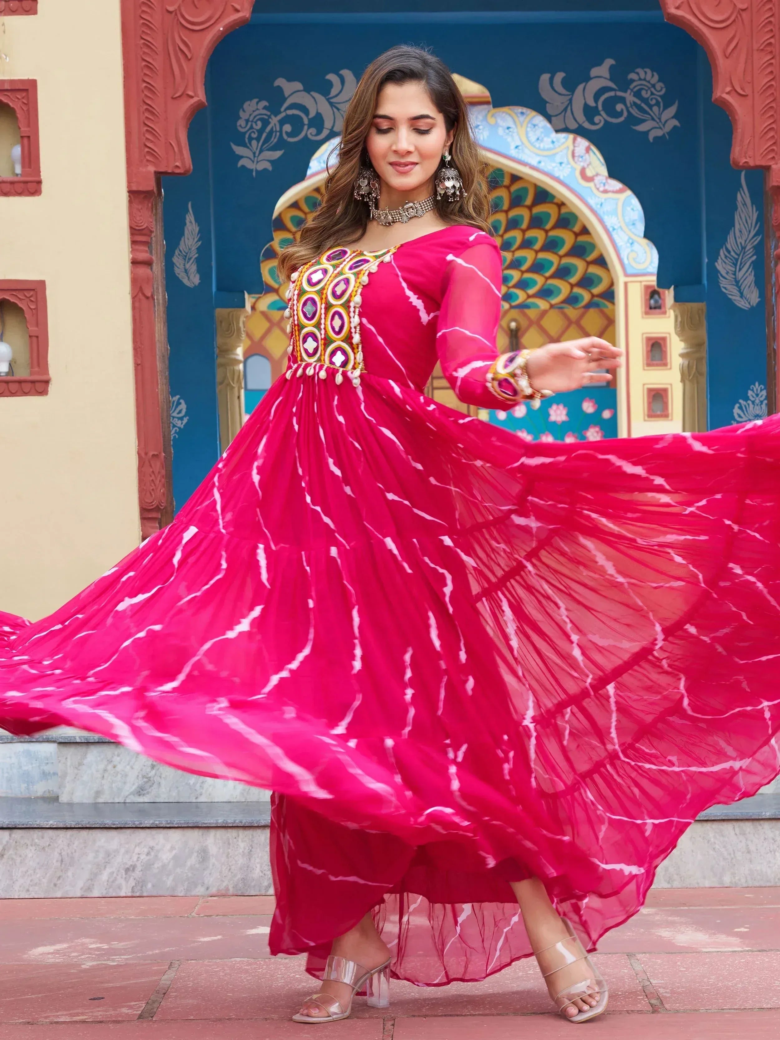 Woman in vibrant pink ethnic gown with mirror work, twirling against colorful Indian architecture