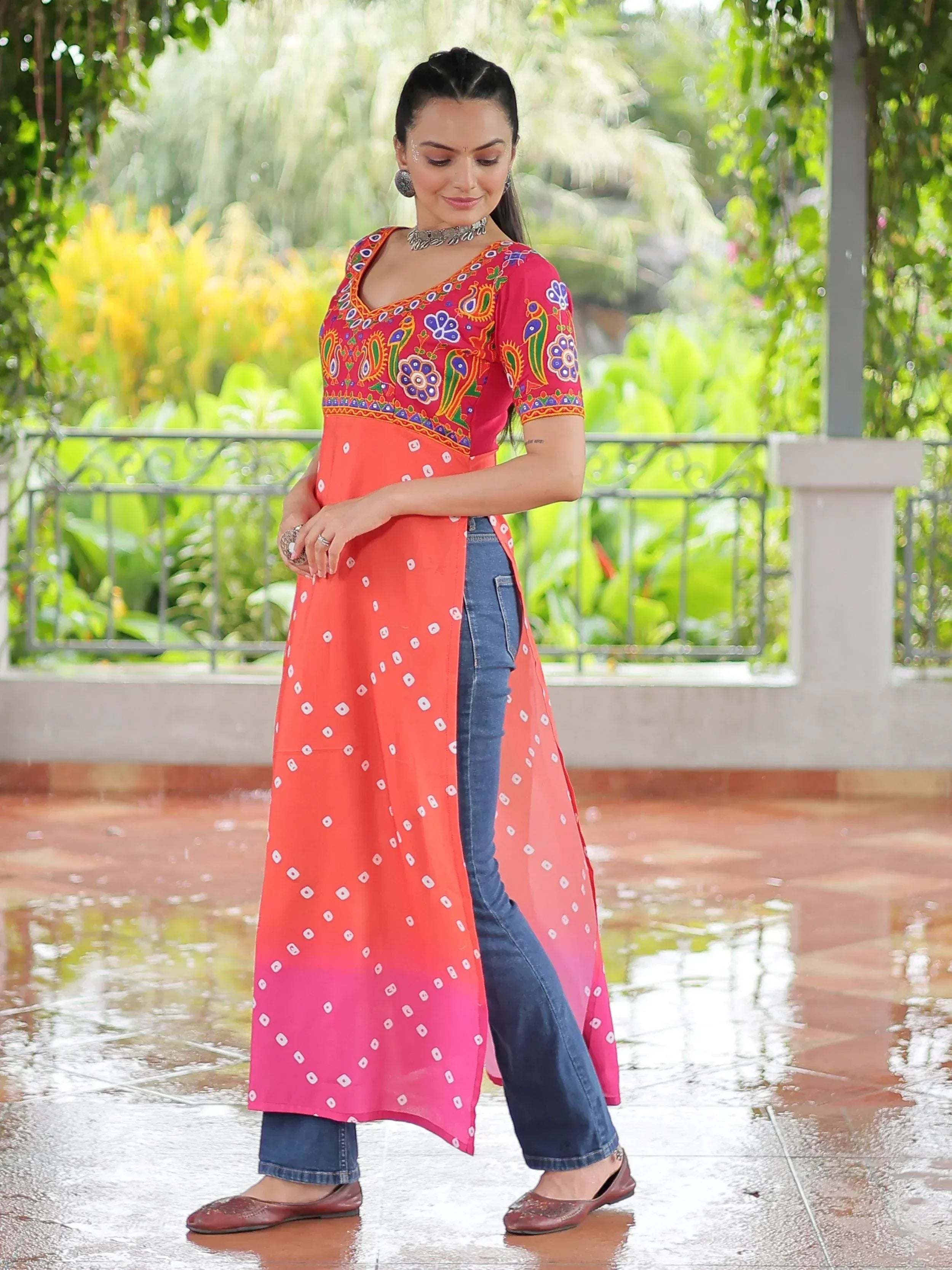 Woman in vibrant ethnic kurti with peacock embroidery, paired with jeans, standing outdoors on wet tiles.