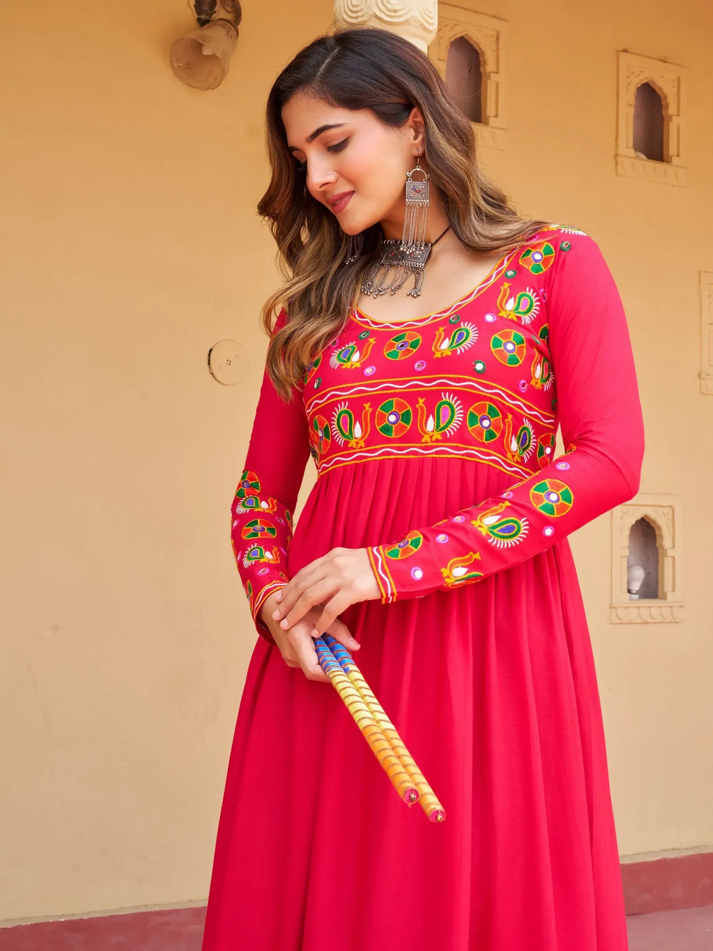 Vibrant red ethnic gown with colorful embroidery, paired with traditional jewelry and dandiya sticks.