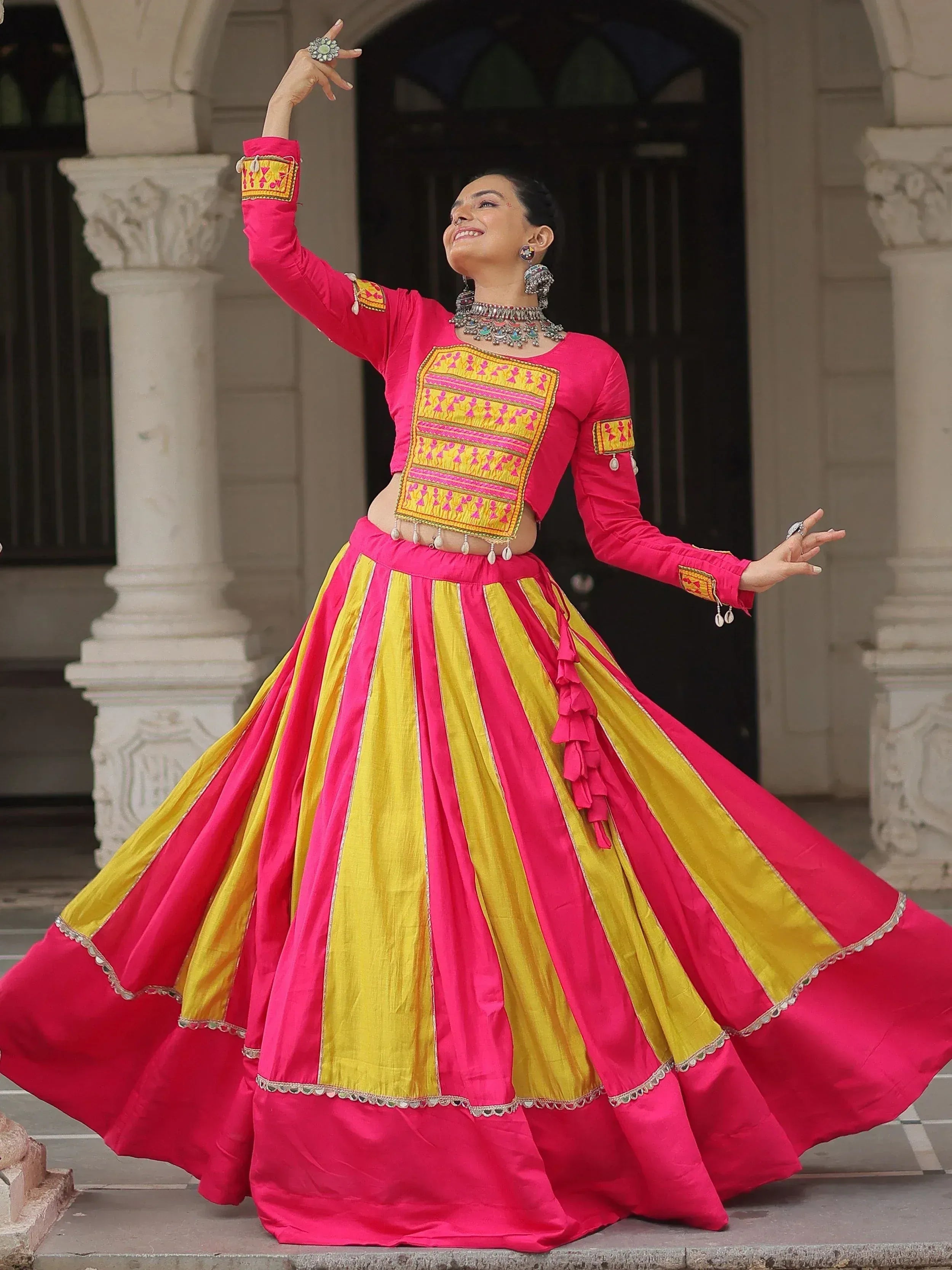 Woman in vibrant pink and yellow Indian ethnic lehenga choli with embroidery, dancing gracefully outdoors.